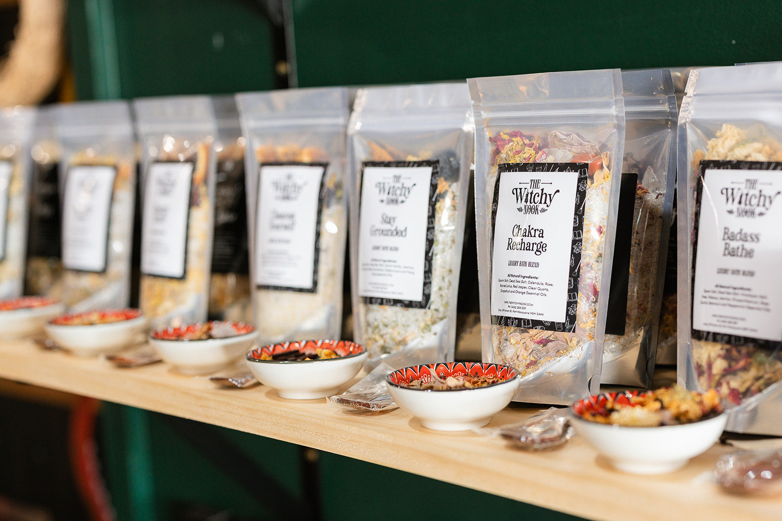 A shelf with a row of The Witchy Nook branded packets of bath salts with small bowls of samples in front 