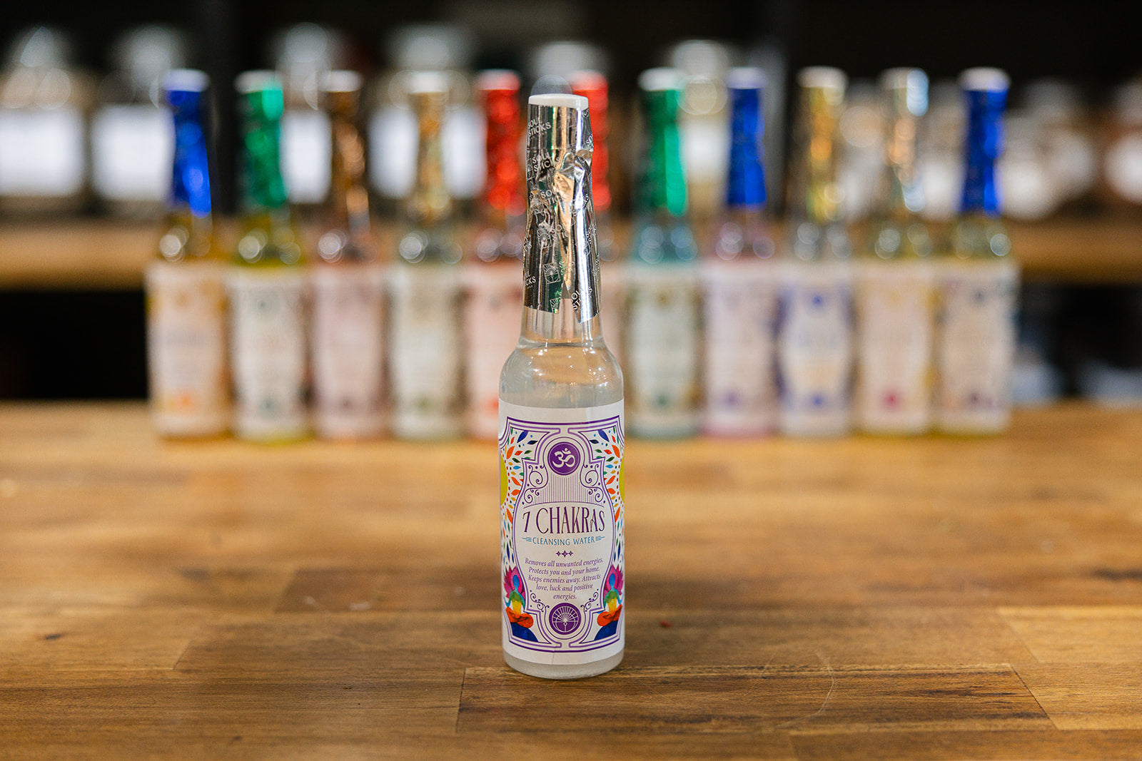 7 Chakras Cleansing Water on a timber background with a collection of cleansing waters displayed in the background.