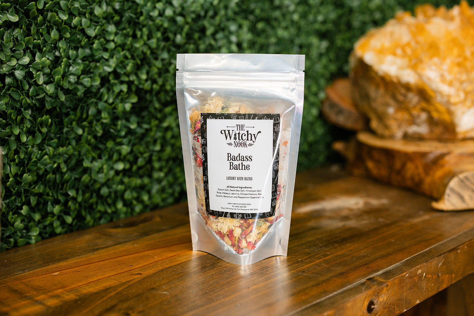 Packaged product labeled 'Badarse Bathe Bath Blend' by 'Witchy Nook' on a wooden surface with greenery in the background.