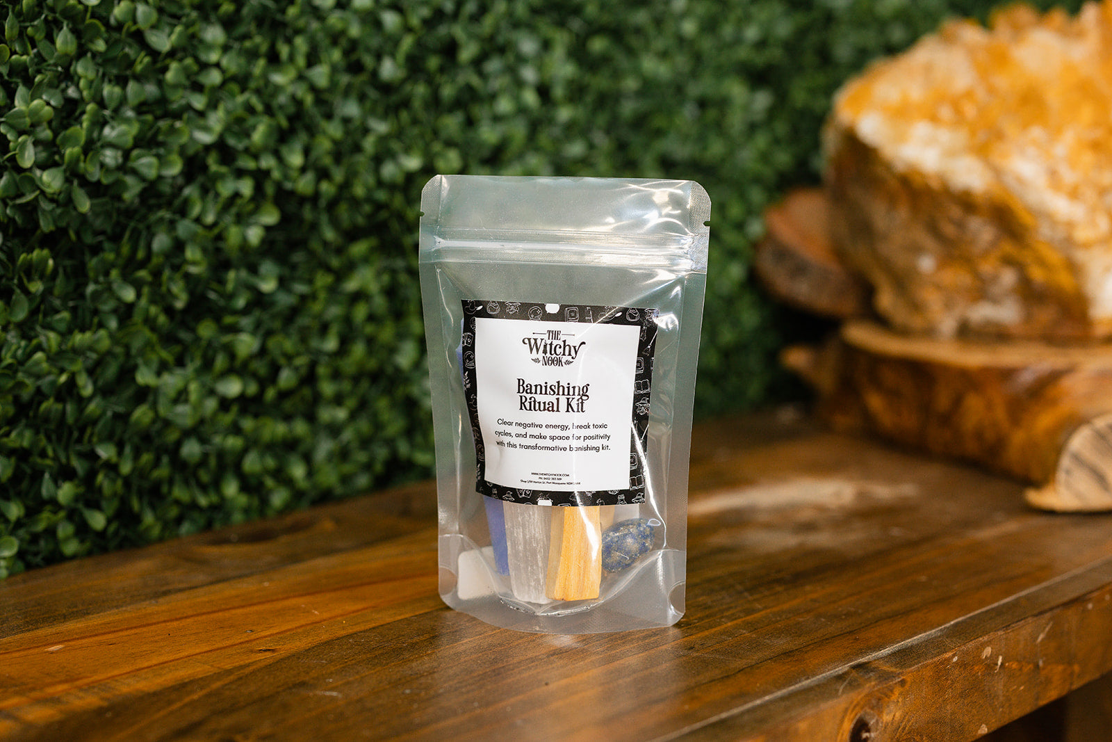 Packaged product labeled 'Banishing Ritual Kit' by 'Witchy Nook' on a wooden surface with greenery in the background.