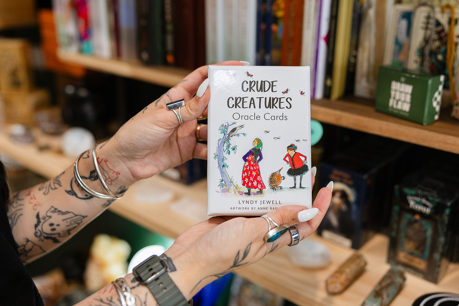 Crude Creatures Oracle cards by Lyndy Jewell held by a person displayed in front of a bookshelf