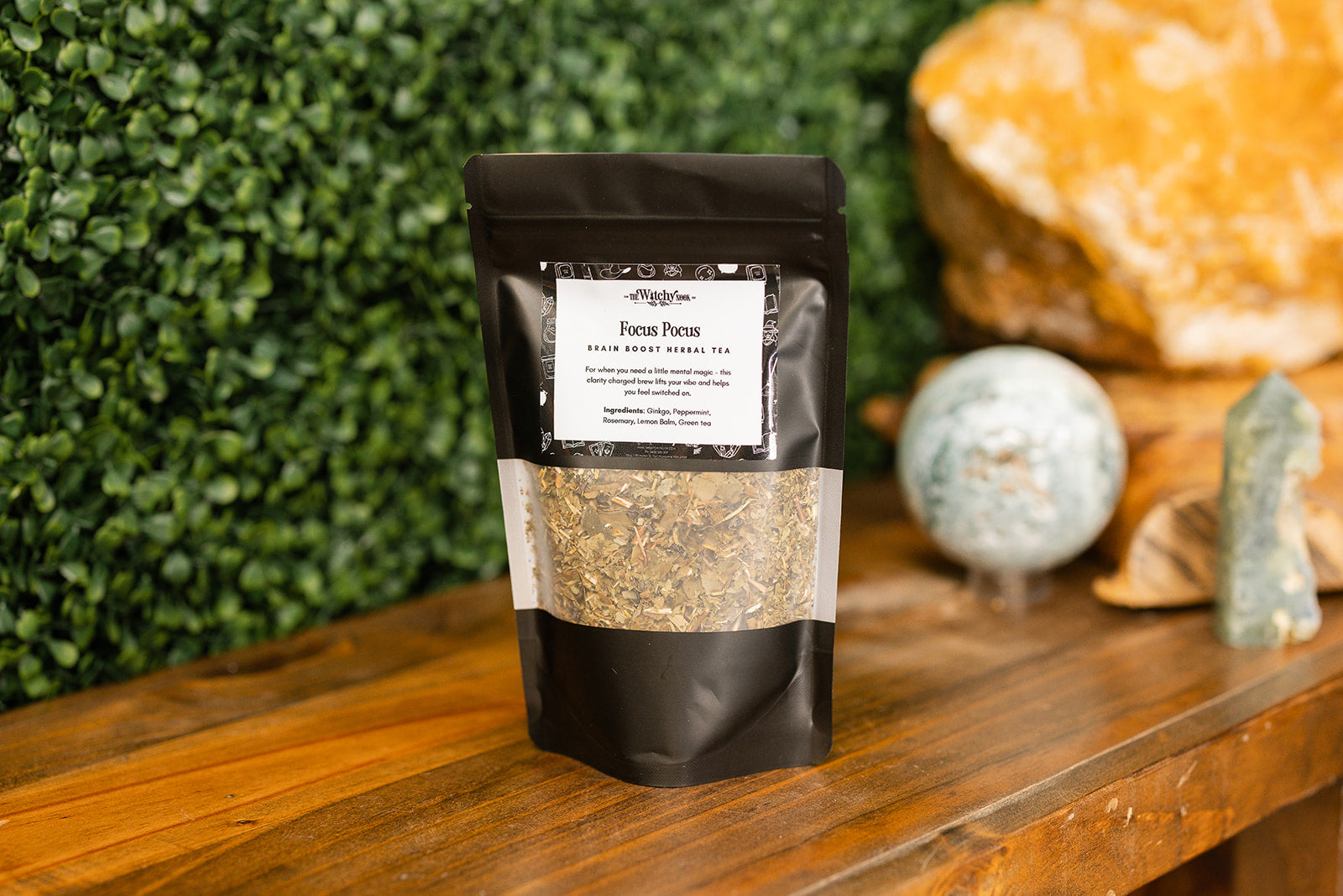 Packaged product labeled 'Focus Pocus Tea Blend' by 'Witchy Nook' on a wooden surface with greenery in the background.