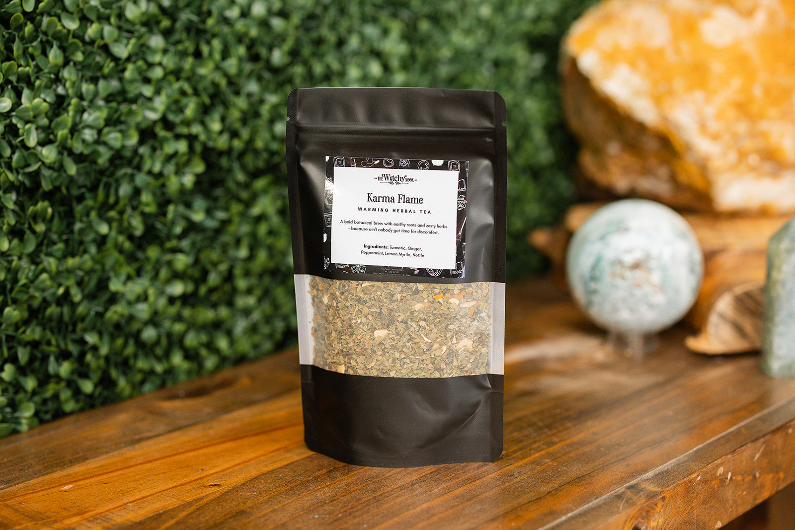 Packaged product labeled 'Karma Flame Tea Blend' by 'Witchy Nook' on a wooden surface with greenery in the background.