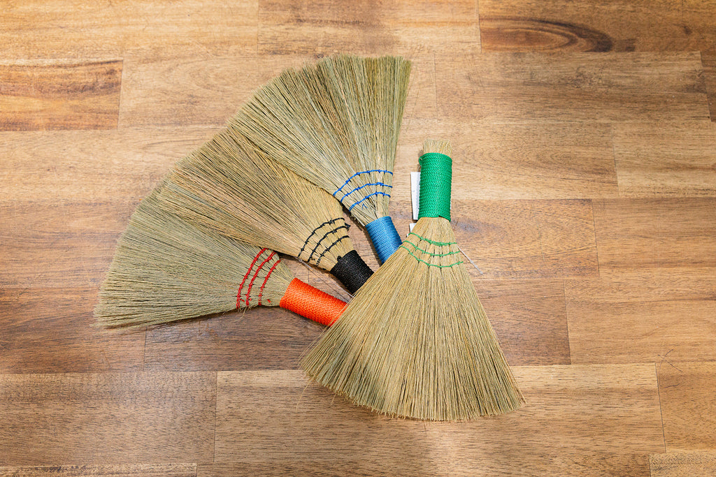 The image displays several Mini Brooms - Assorted Colours made from natural fibres displayed on a timber background