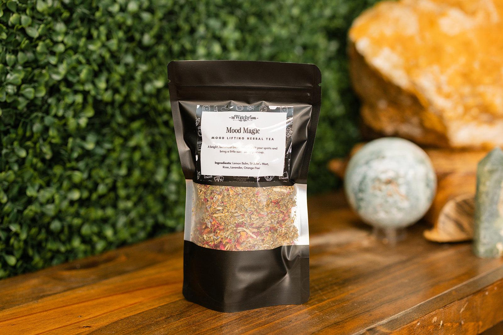 Packaged product labeled 'Mood Magic Tea Blend' by 'Witchy Nook' on a wooden surface with greenery in the background.