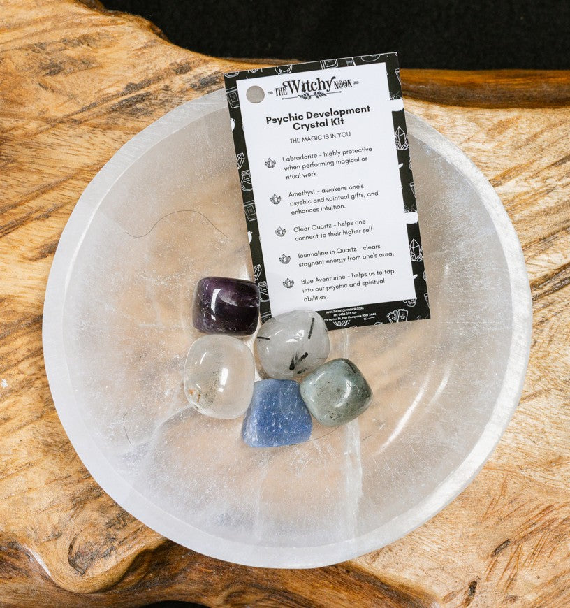 Packaged product labeled 'Psychic Development Tumble Kit' by 'Witchy Nook' on a wooden surface in the background with products displayed in a bowl.