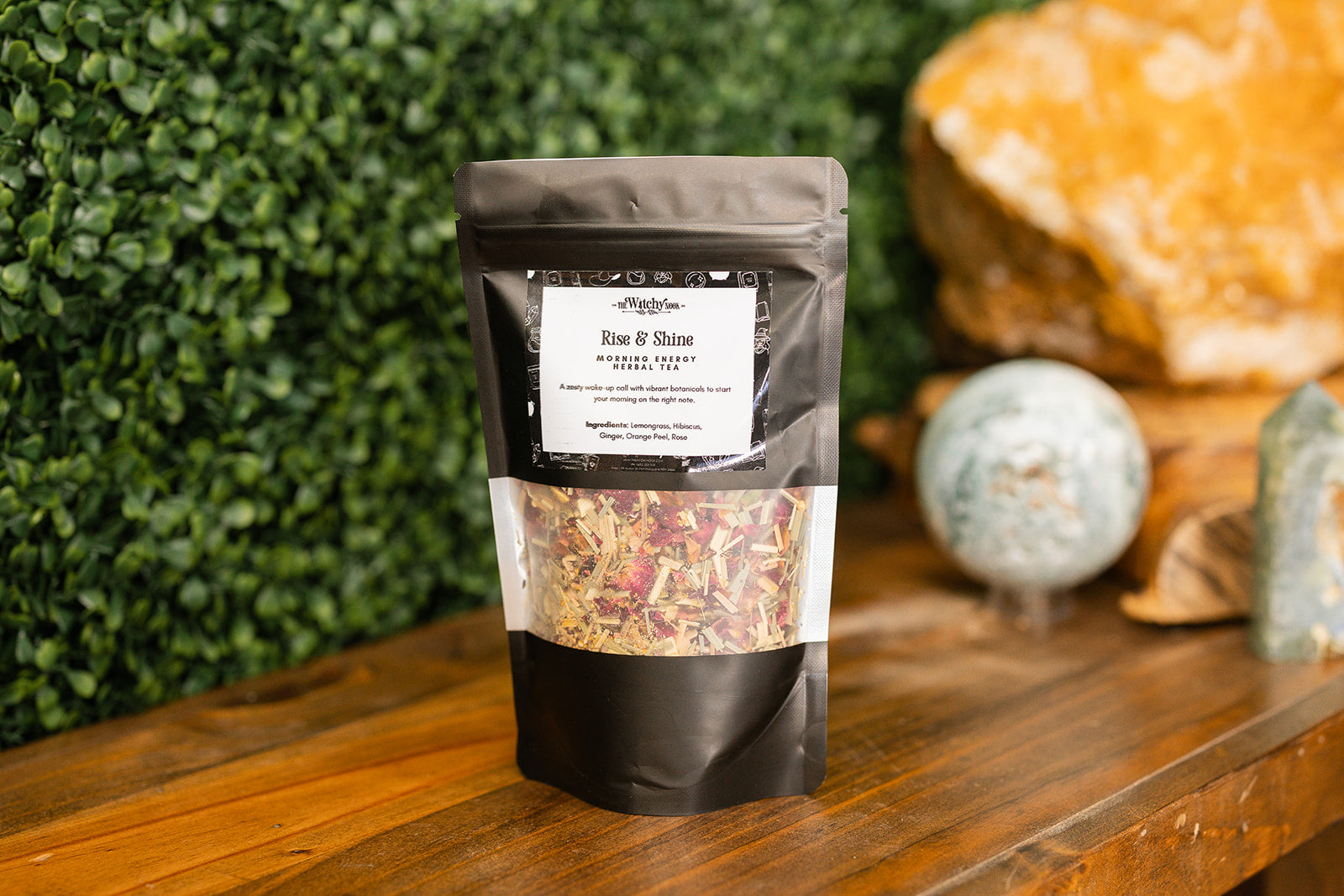 Packaged product labeled 'Rise and Shine Tea Blend' by 'Witchy Nook' on a wooden surface with greenery in the background.