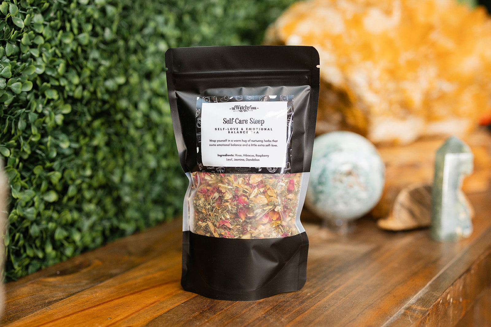 Packaged product labeled 'Self-Care Steep Tea Blend' by 'Witchy Nook' on a wooden surface with greenery in the background.