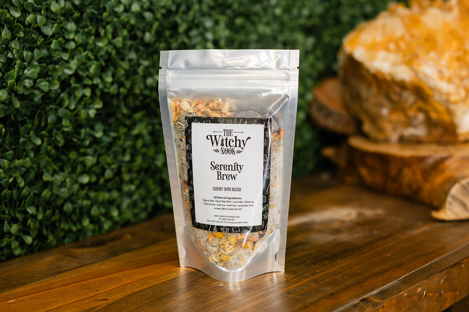 Packaged product labeled 'Serenity Brew Bath Blend' by 'Witchy Nook' on a wooden surface with greenery in the background.