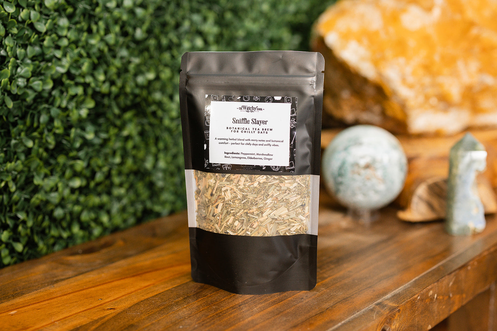 Packaged product labeled 'Sniffle Slayer Tea Blends' by 'Witchy Nook' on a wooden surface with greenery in the background.