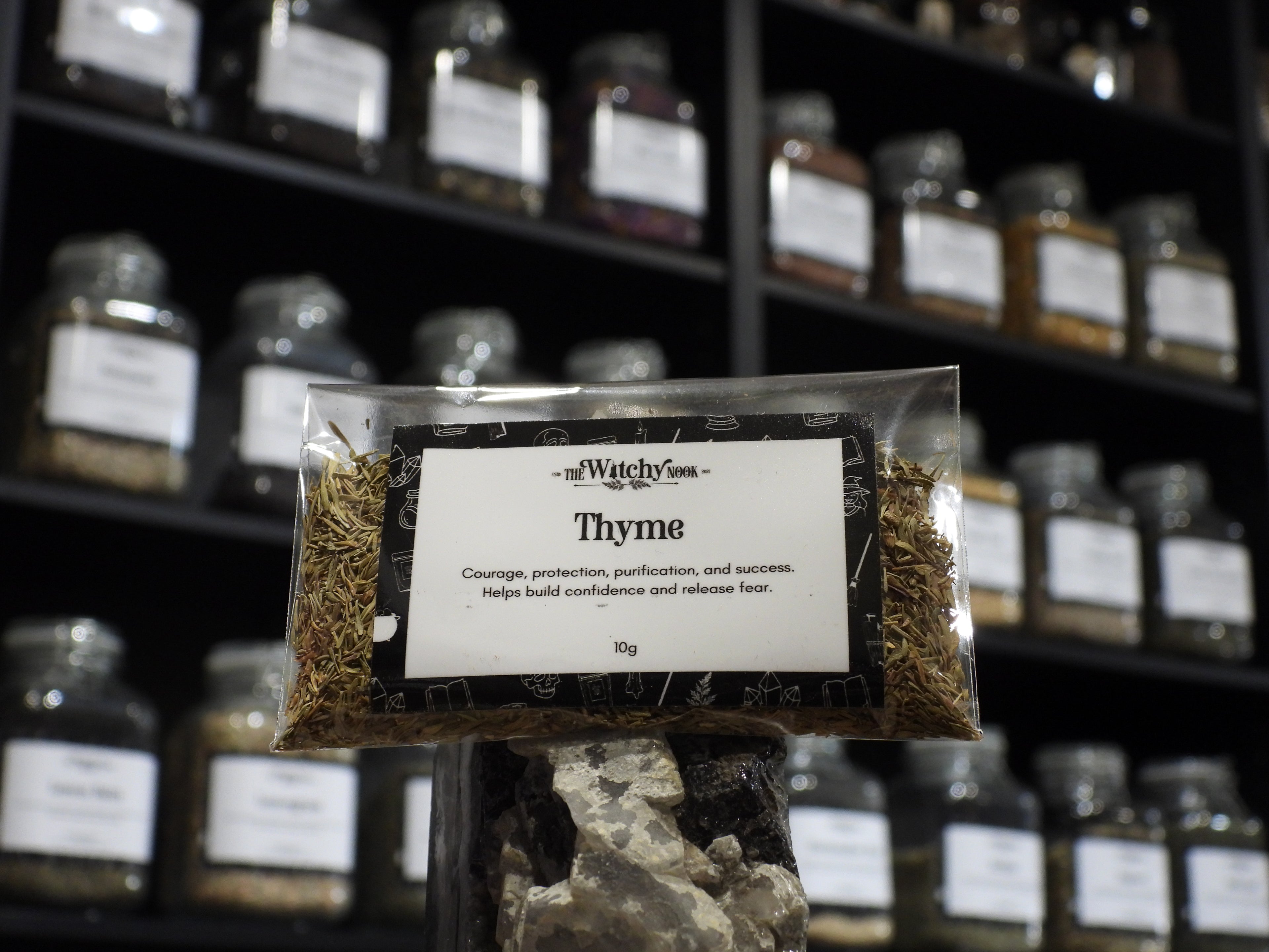 Packaged product labelled ‘Thyme' by 'Witchy Nook' sitting on a crystal with an apothecary of herb jars blurred in the background.