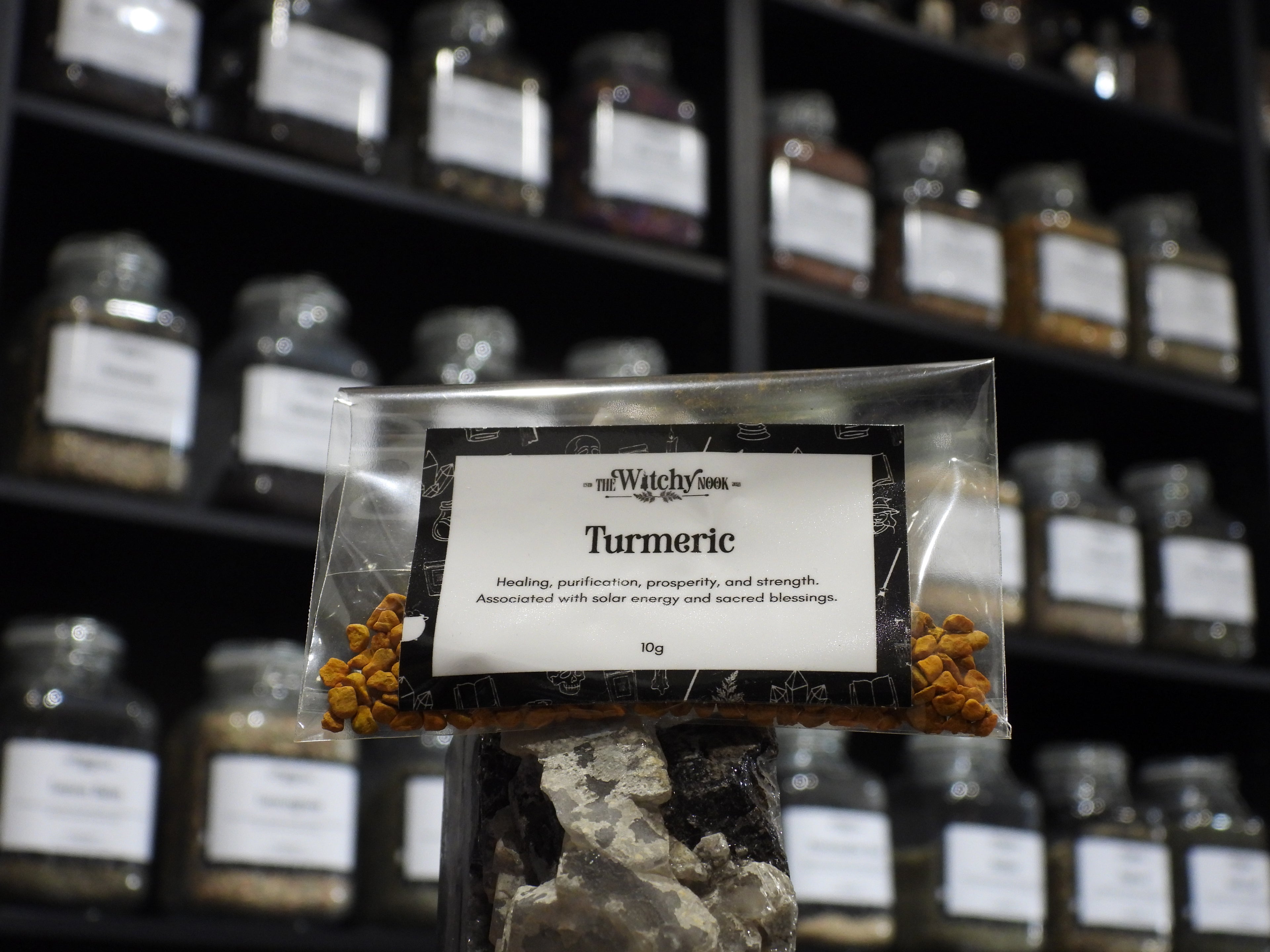 Packaged product labelled ‘Turmeric' by 'Witchy Nook' sitting on a crystal with an apothecary of herb jars blurred in the background.