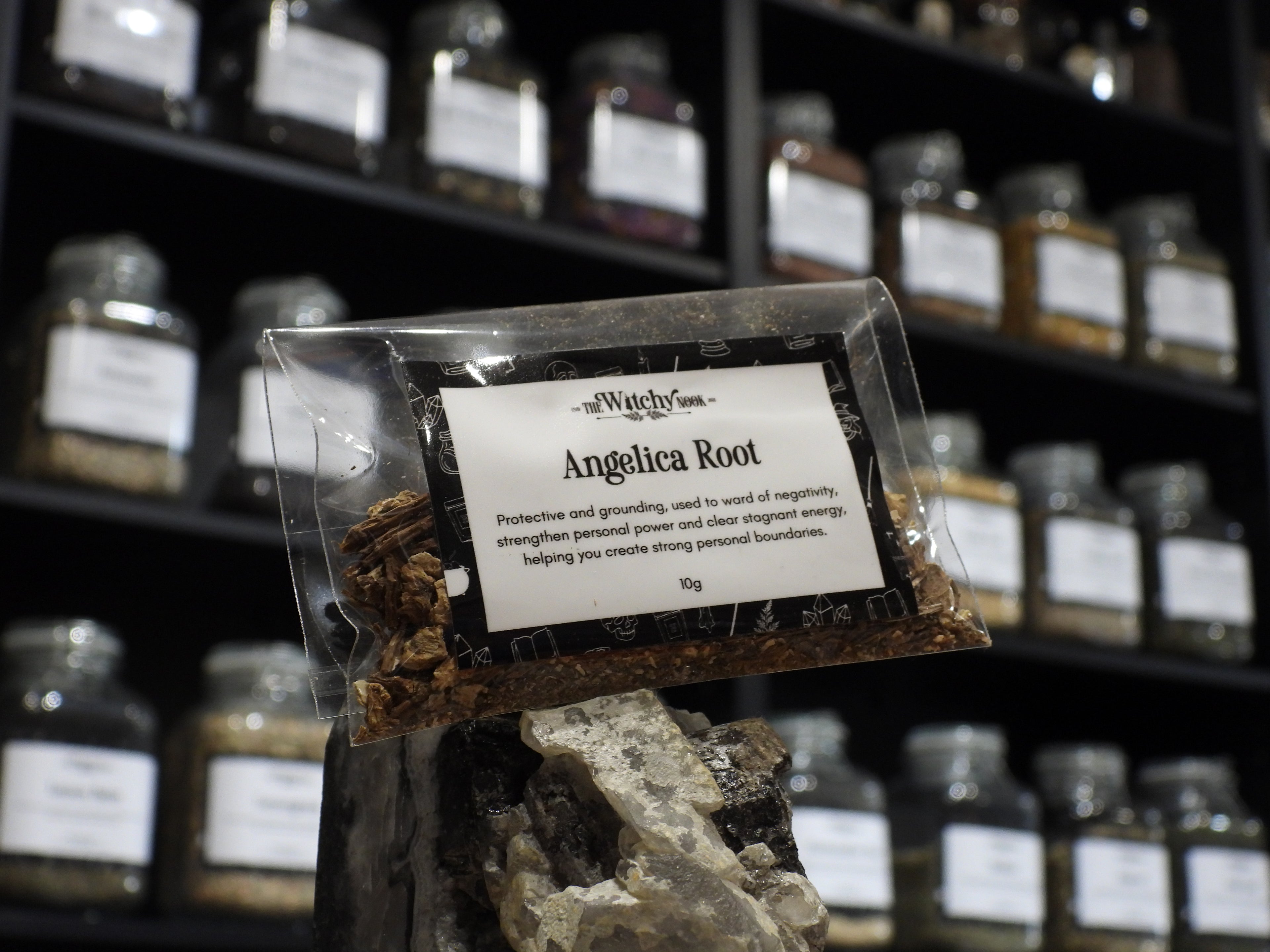 Packaged product labelled ‘Angelica Root' by 'Witchy Nook' sitting on a crystal with an apothecary of herb jars blurred in the background.