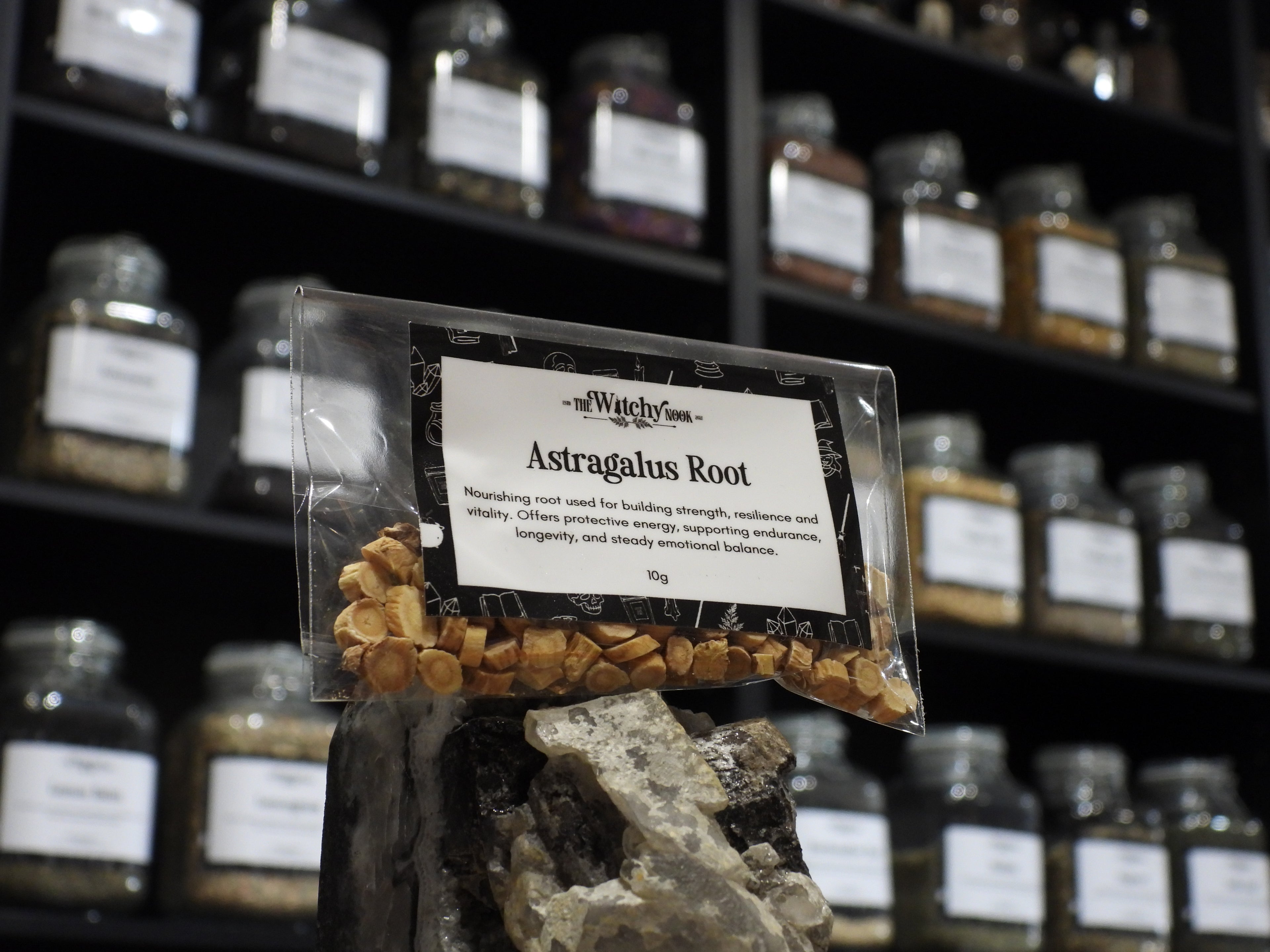 Packaged product labelled ‘Astragalus Root' by 'Witchy Nook' sitting on a crystal with an apothecary of herb jars blurred in the background.