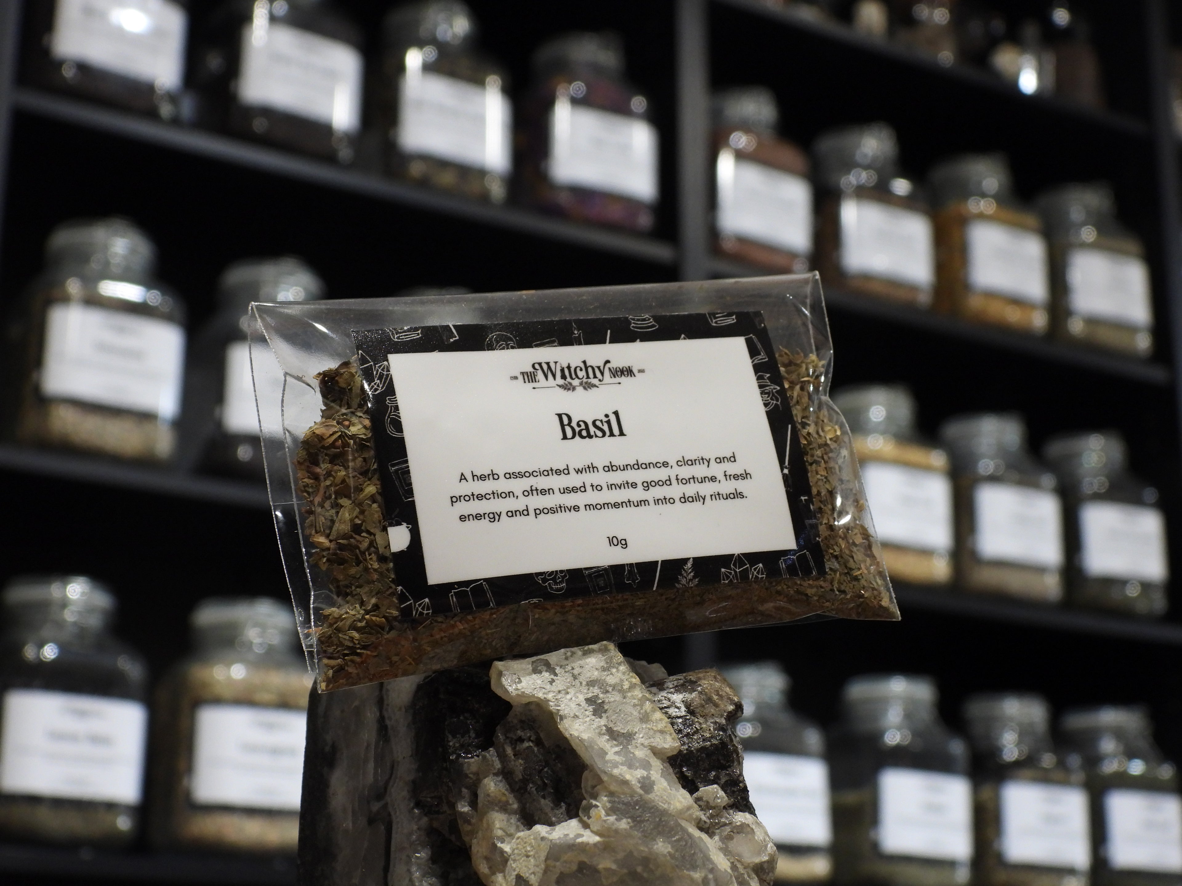 Packaged product labelled ‘Basil’ by 'The Witchy Nook' sitting on a crystal with an apothecary of herb jars blurred in the background.