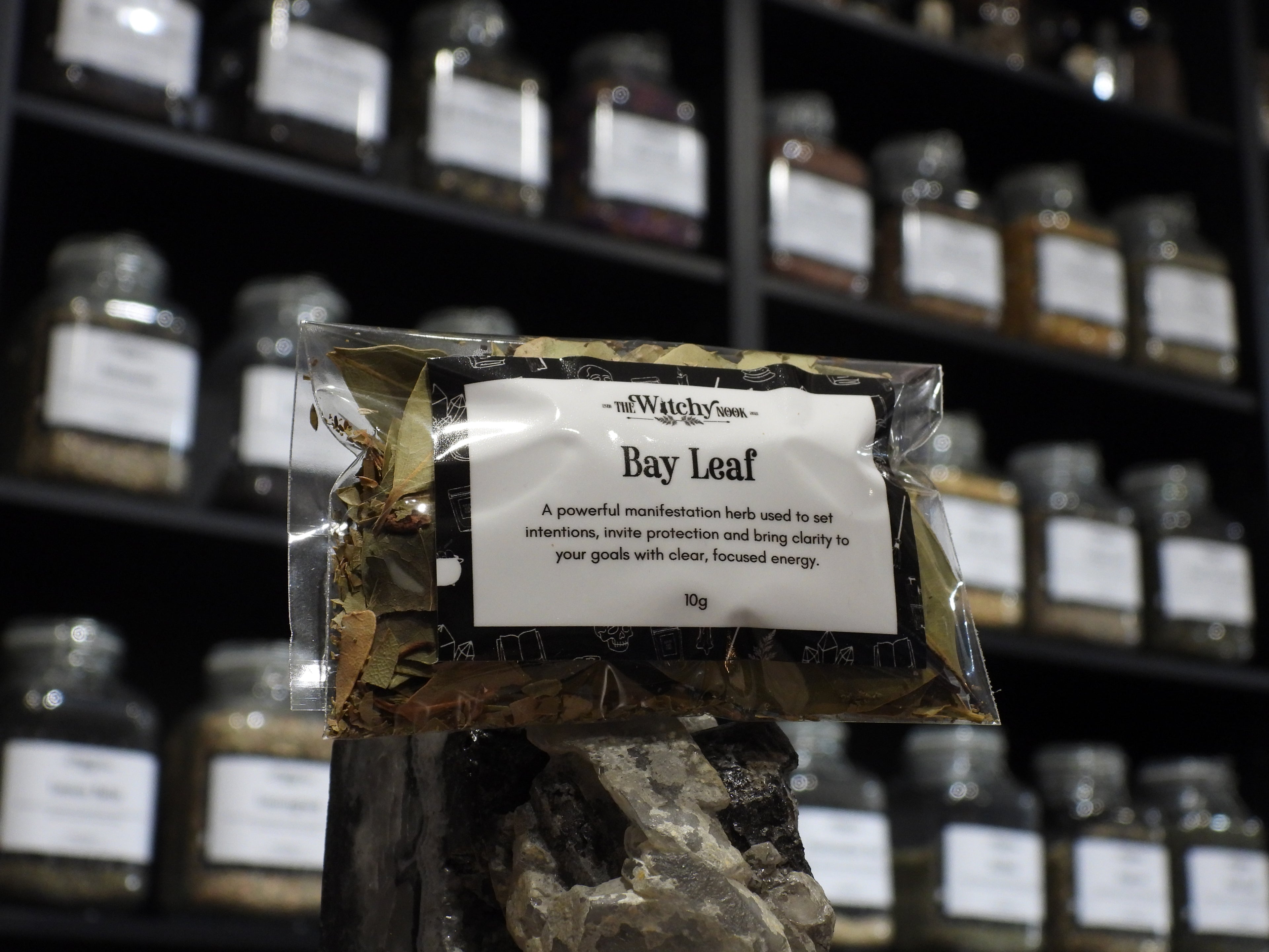 Packaged product labelled ‘Bay Leaf’ by 'The Witchy Nook' sitting on a crystal with an apothecary of herb jars blurred in the background.