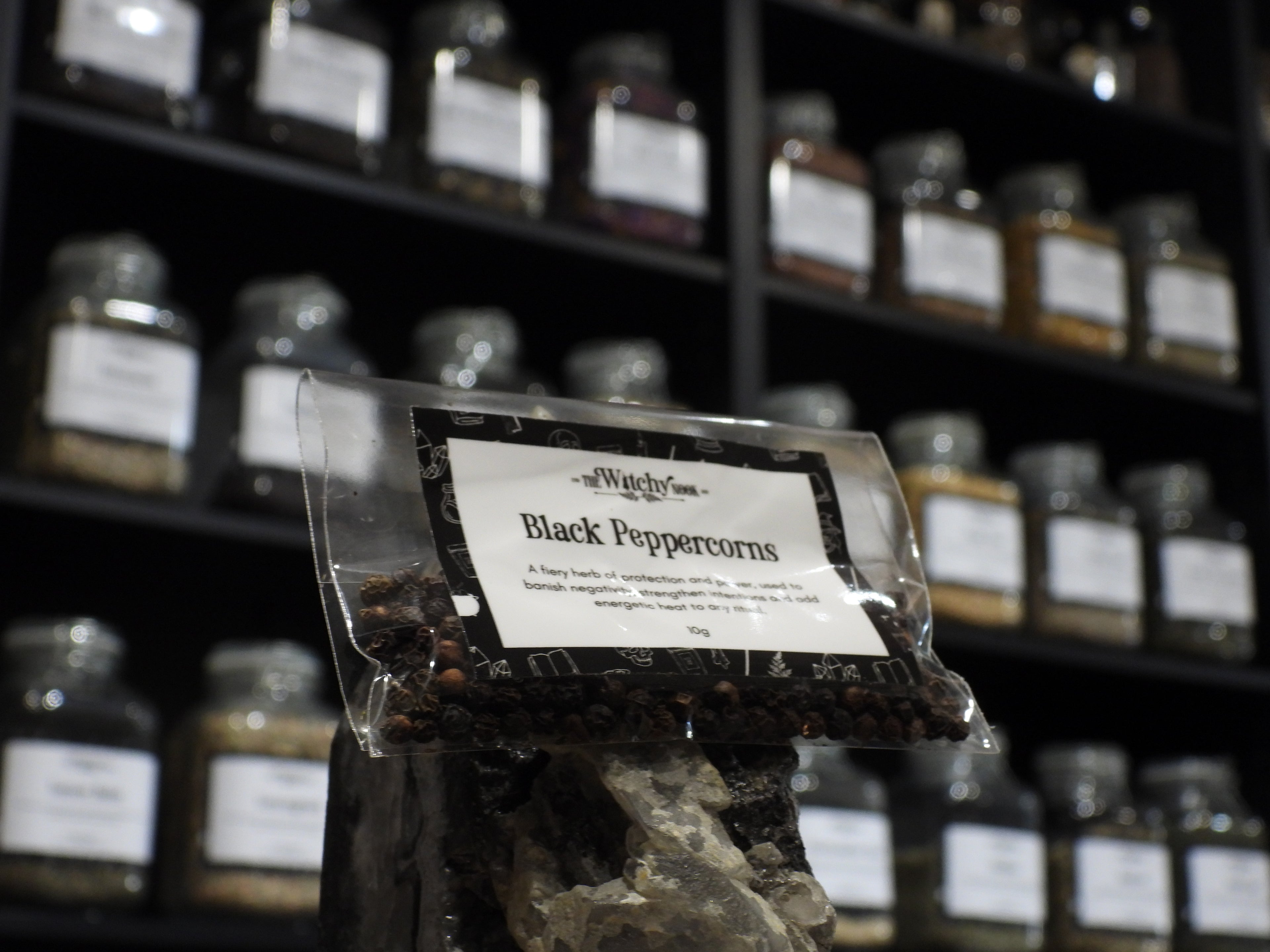 Packaged product labelled ‘Black Peppercorns' by 'Witchy Nook' sitting on a crystal with an apothecary of herb jars blurred in the background.