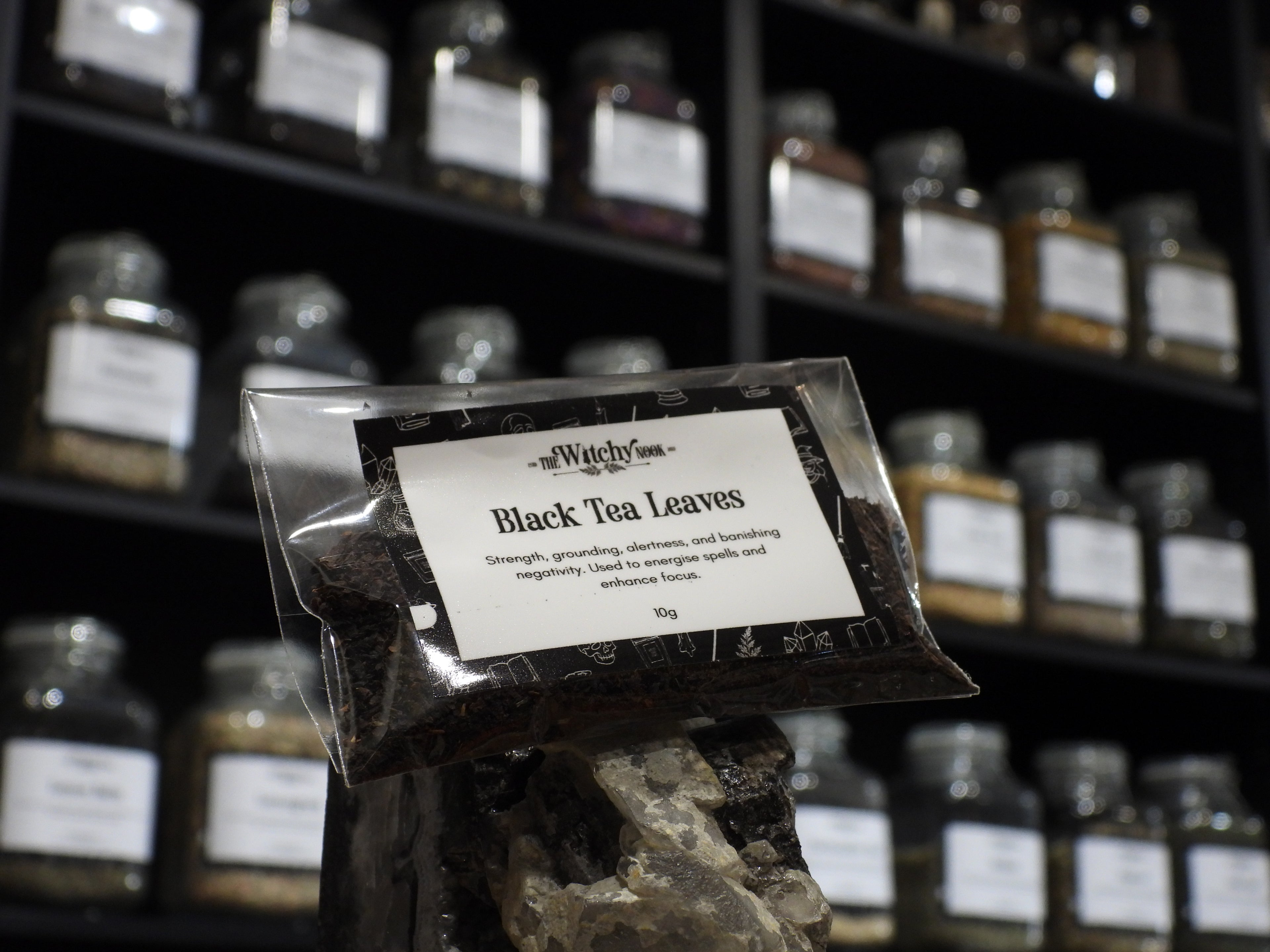 Packaged product labelled ‘Black Tea Leaves' by 'Witchy Nook' sitting on a crystal with an apothecary of herb jars blurred in the background.