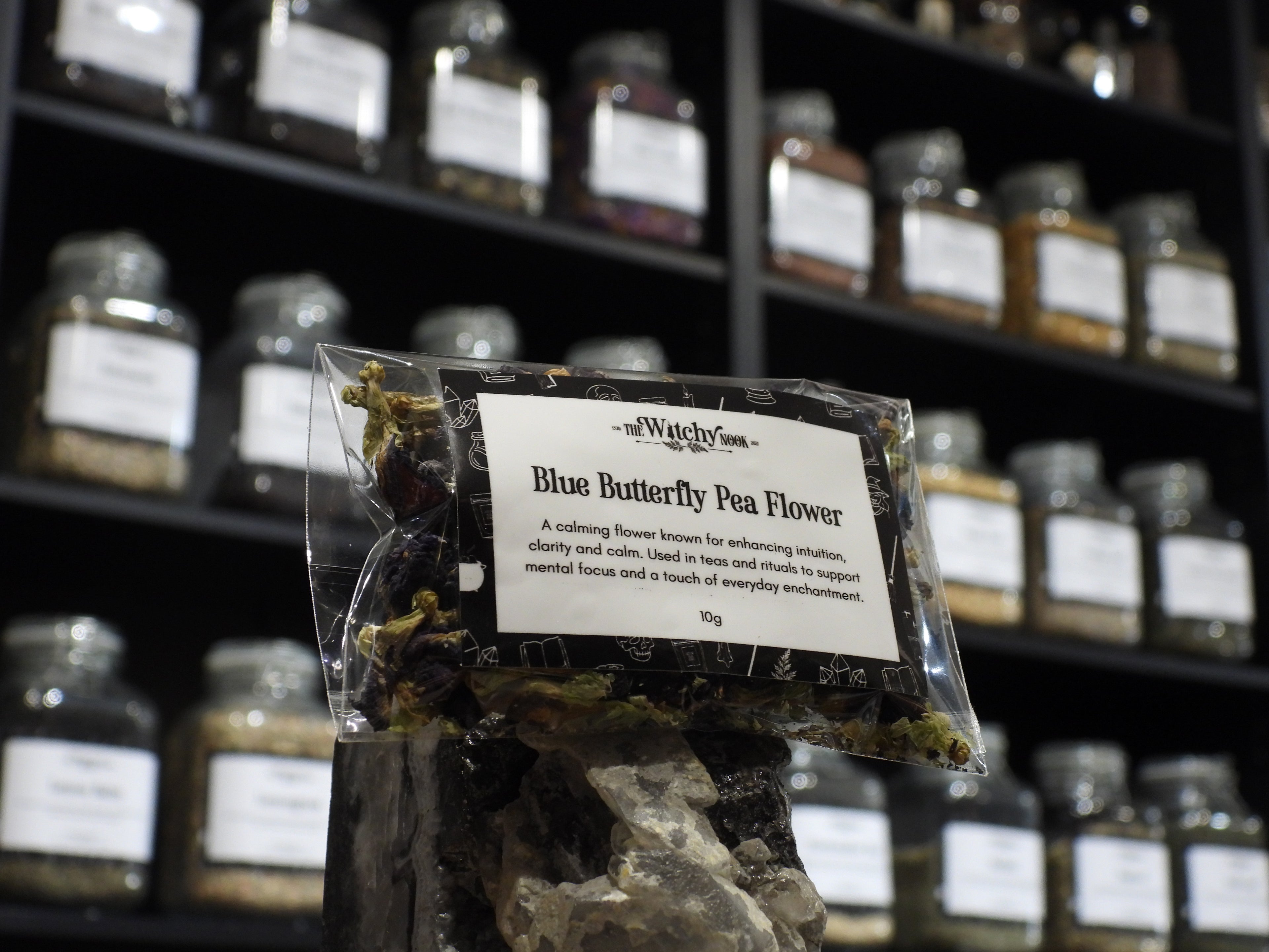 Packaged product labelled ‘Blue Butterfly Pea Flower' by 'Witchy Nook' sitting on a crystal with an apothecary of herb jars blurred in the background.