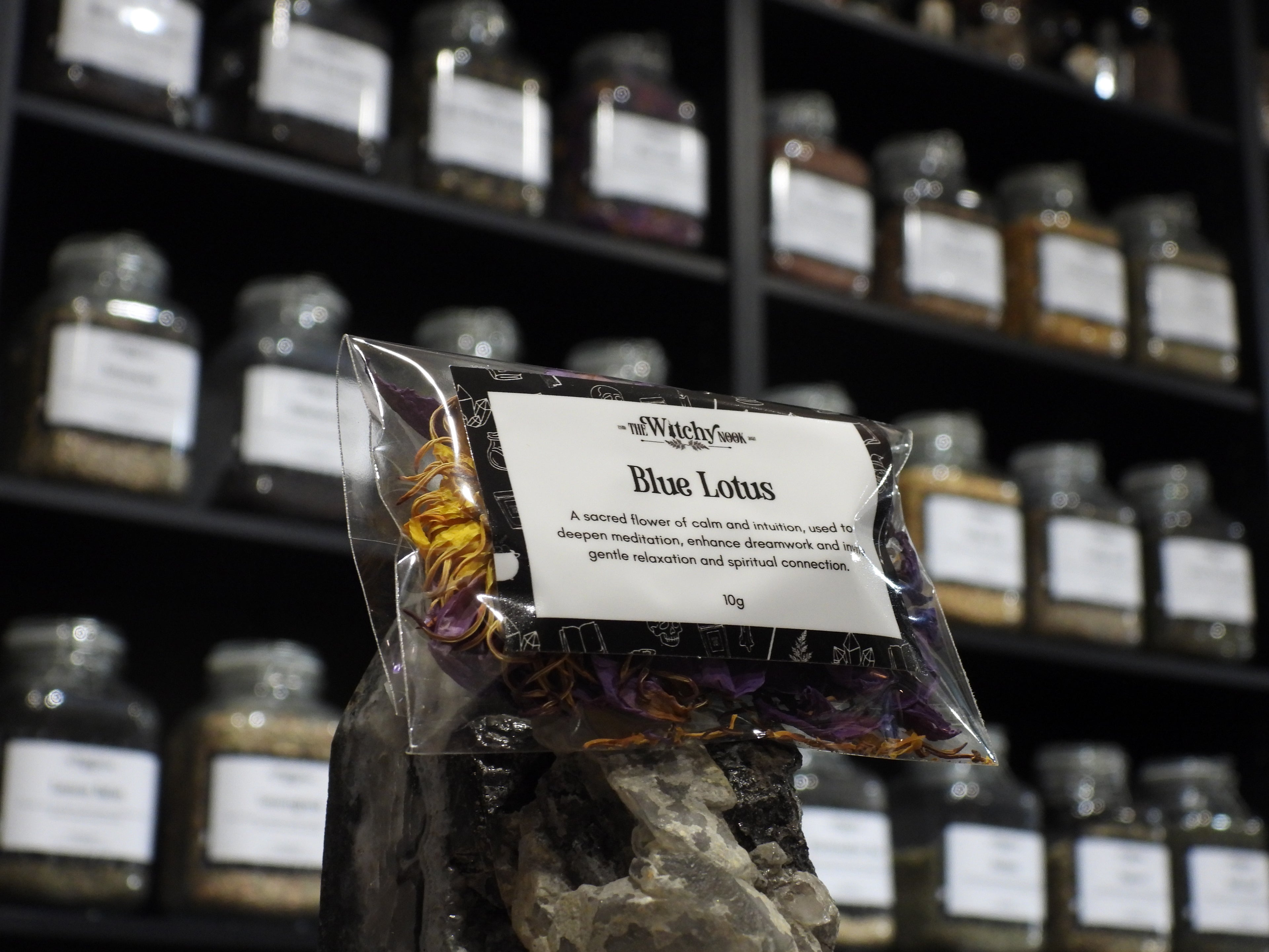 Packaged product labelled ‘Blue Lotus' by 'Witchy Nook' sitting on a crystal with an apothecary of herb jars blurred in the background.