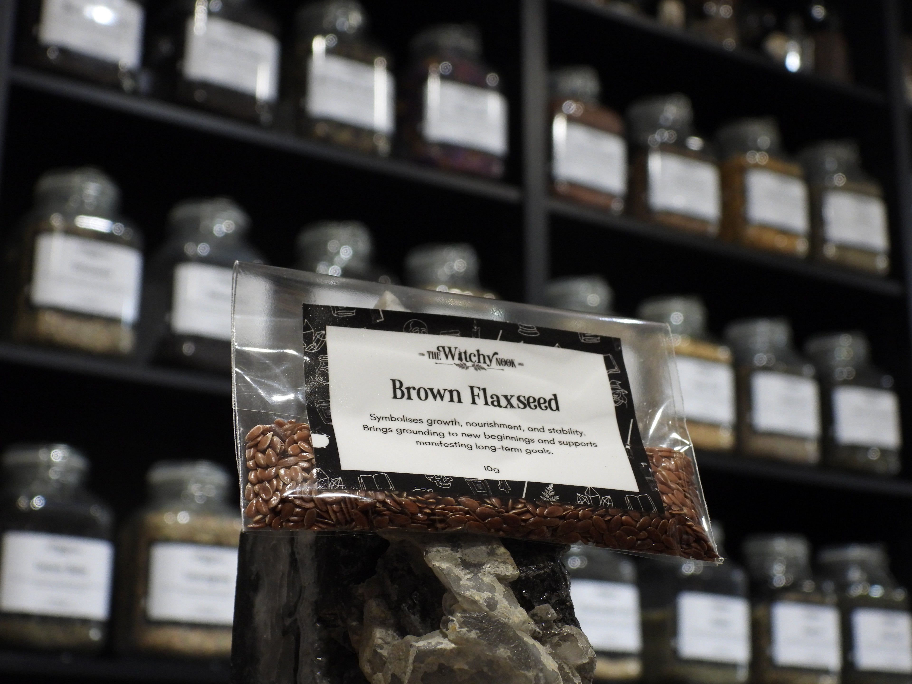 Packaged product labelled ‘Brown Flaxseed' by 'Witchy Nook' sitting on a crystal with an apothecary of herb jars blurred in the background.