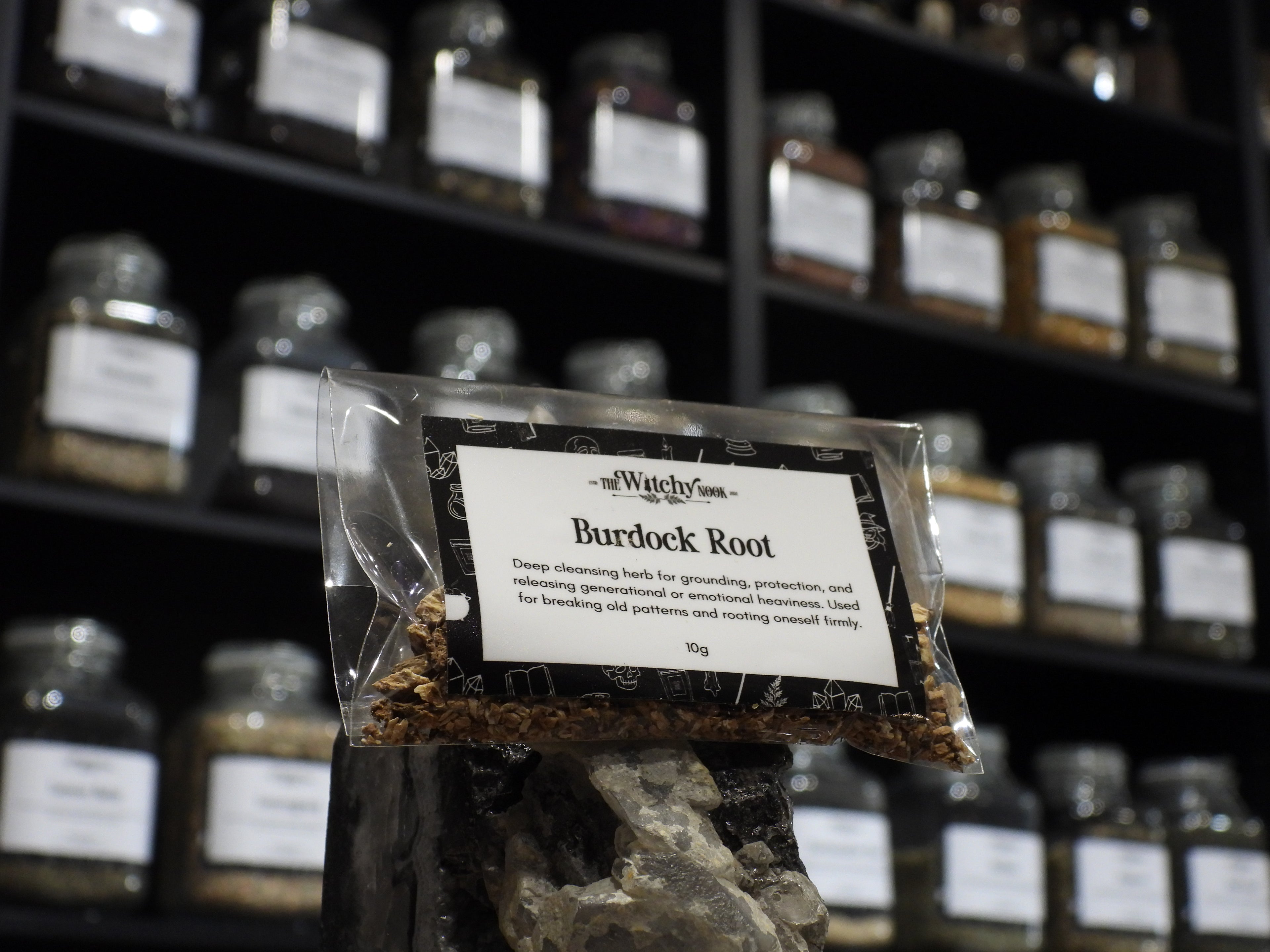 Packaged product labelled ‘Burdock Root' by 'Witchy Nook' sitting on a crystal with an apothecary of herb jars blurred in the background.