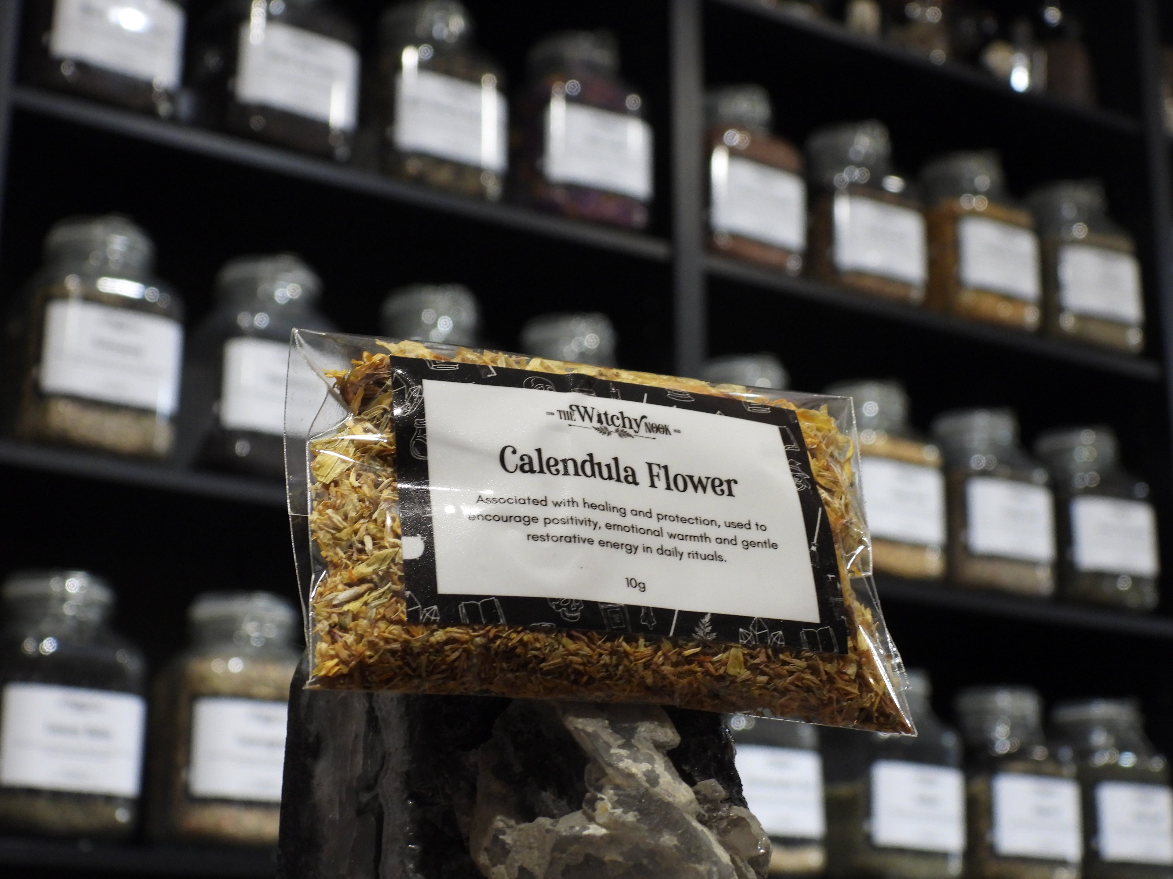 Packaged product labelled ‘Calendula Flower' by 'Witchy Nook' sitting on a crystal with an apothecary of herb jars blurred in the background.