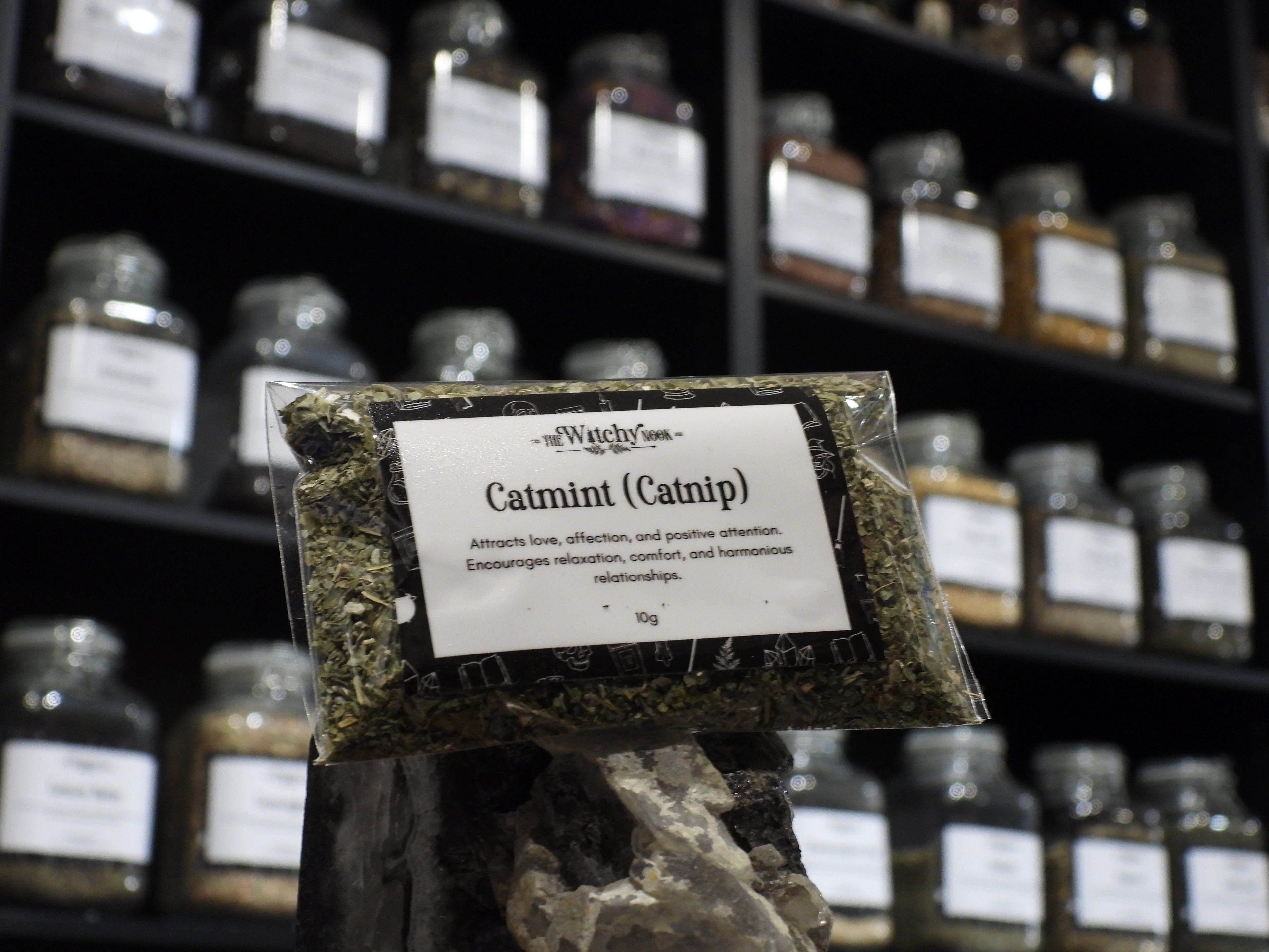 Packaged product labelled ‘Catmint (Catnip)' by 'Witchy Nook' sitting on a crystal with an apothecary of herb jars blurred in the background.