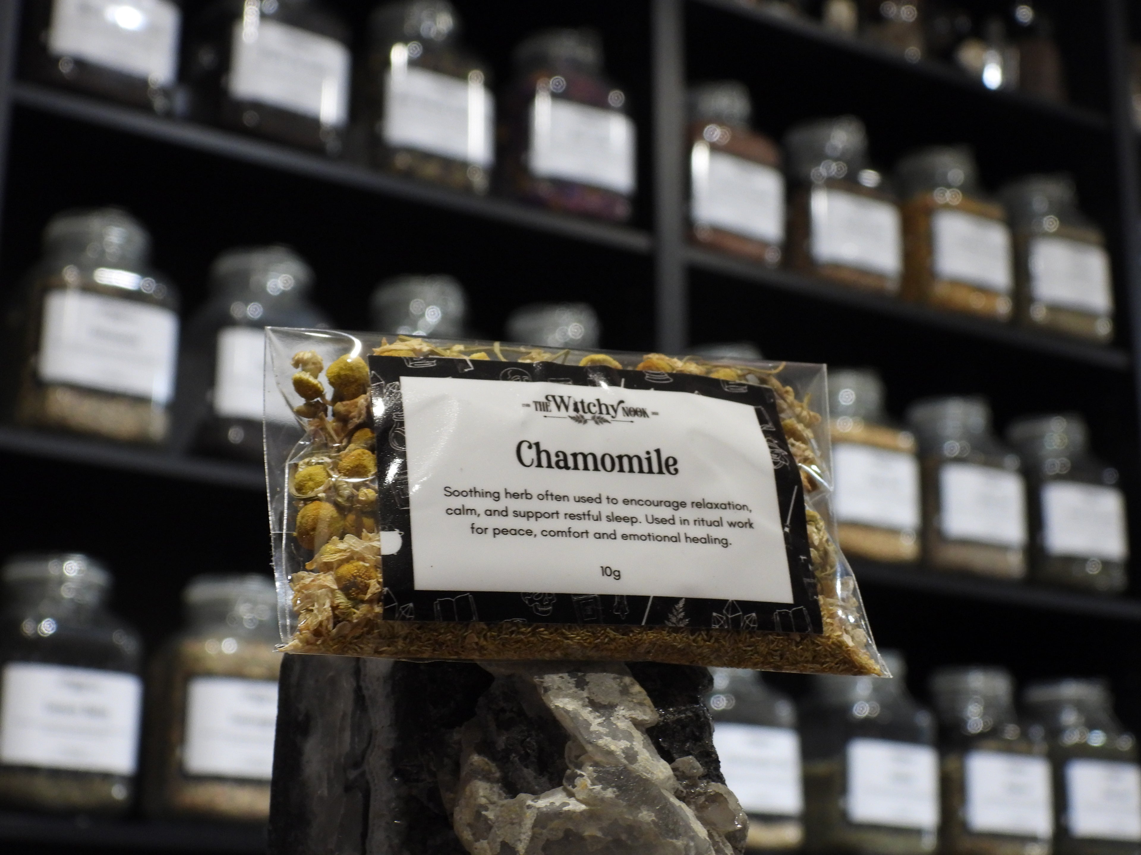 Packaged product labelled ‘Chamomile' by 'Witchy Nook' sitting on a crystal with an apothecary of herb jars blurred in the background.