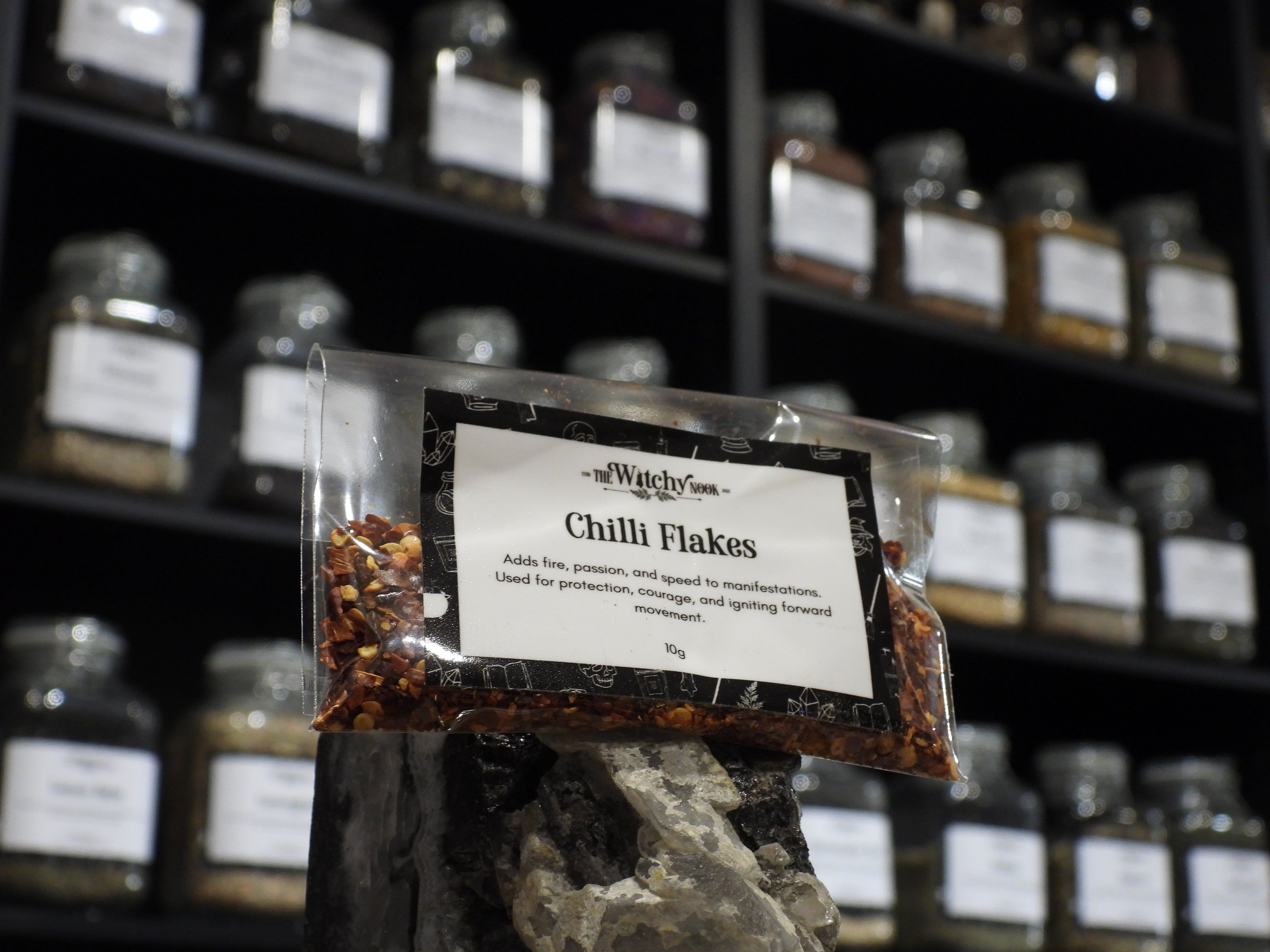 Packaged product labelled ‘Chilli Flakes' by 'Witchy Nook' sitting on a crystal with an apothecary of herb jars blurred in the background.