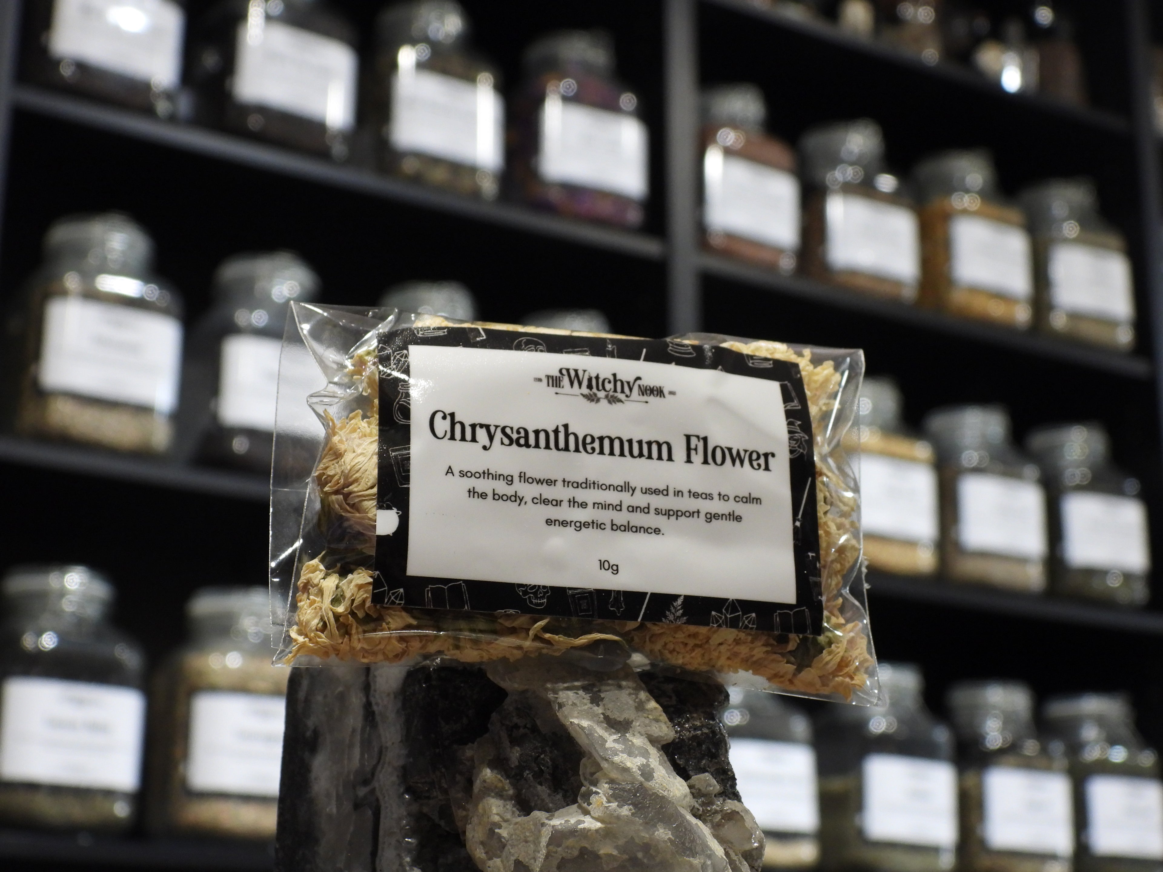 Packaged product labelled ‘Chrysanthemum Flower' by 'Witchy Nook' sitting on a crystal with an apothecary of herb jars blurred in the background.