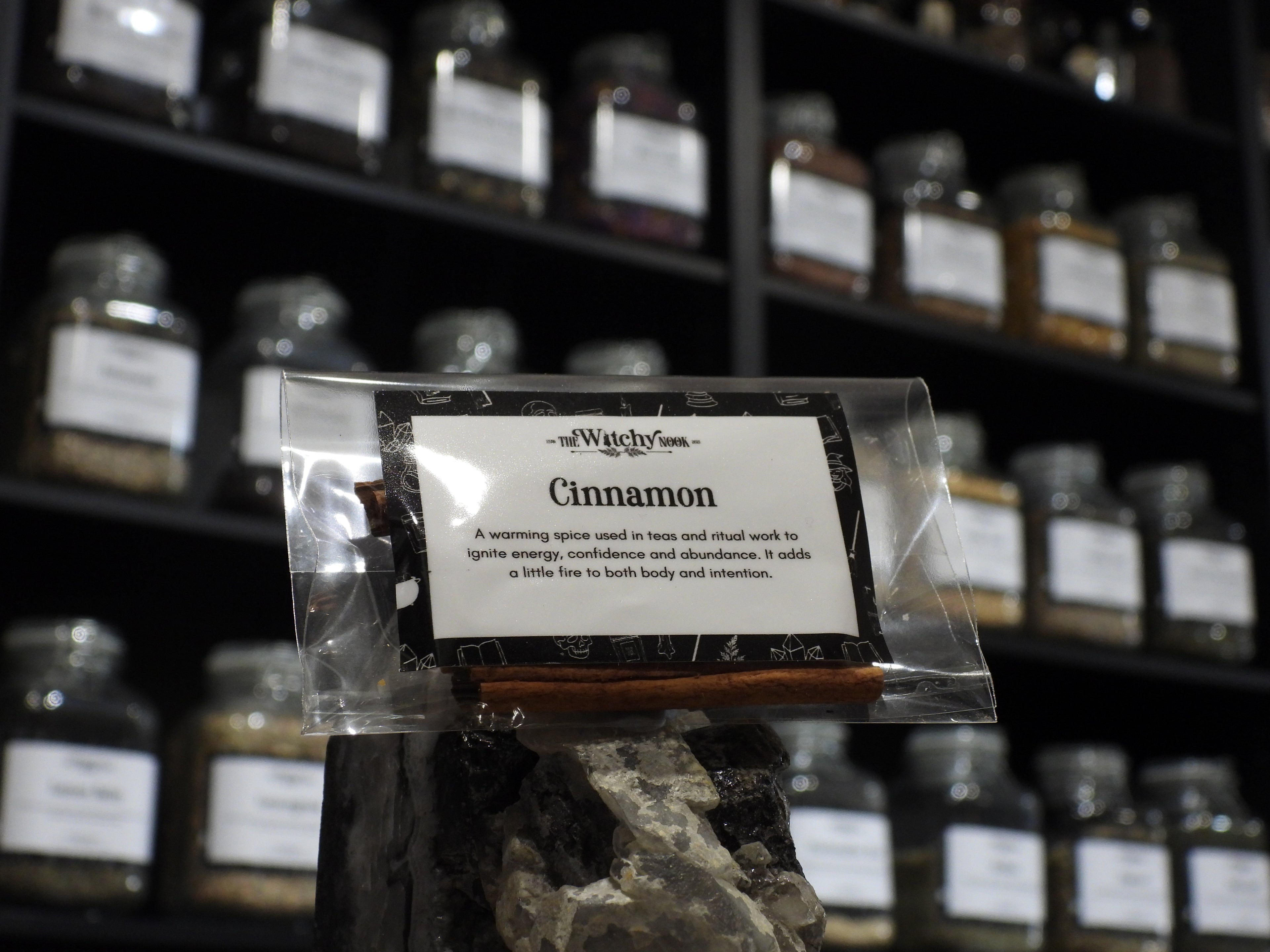 Packaged product labelled ‘Cinnamon' by 'Witchy Nook' sitting on a crystal with an apothecary of herb jars blurred in the background.