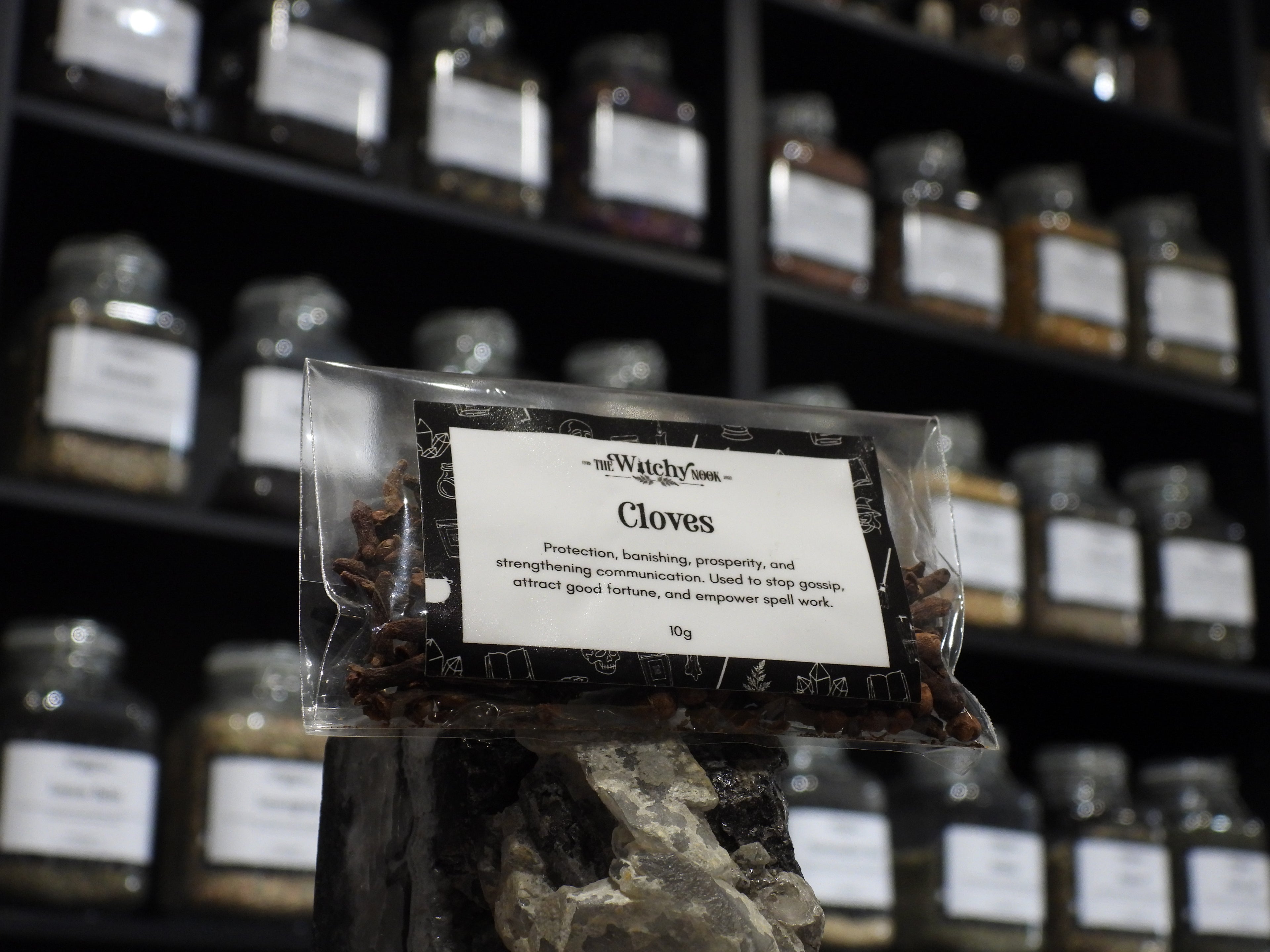 Packaged product labelled ‘Cloves’ by 'The Witchy Nook' sitting on a crystal with an apothecary of herb jars blurred in the background.