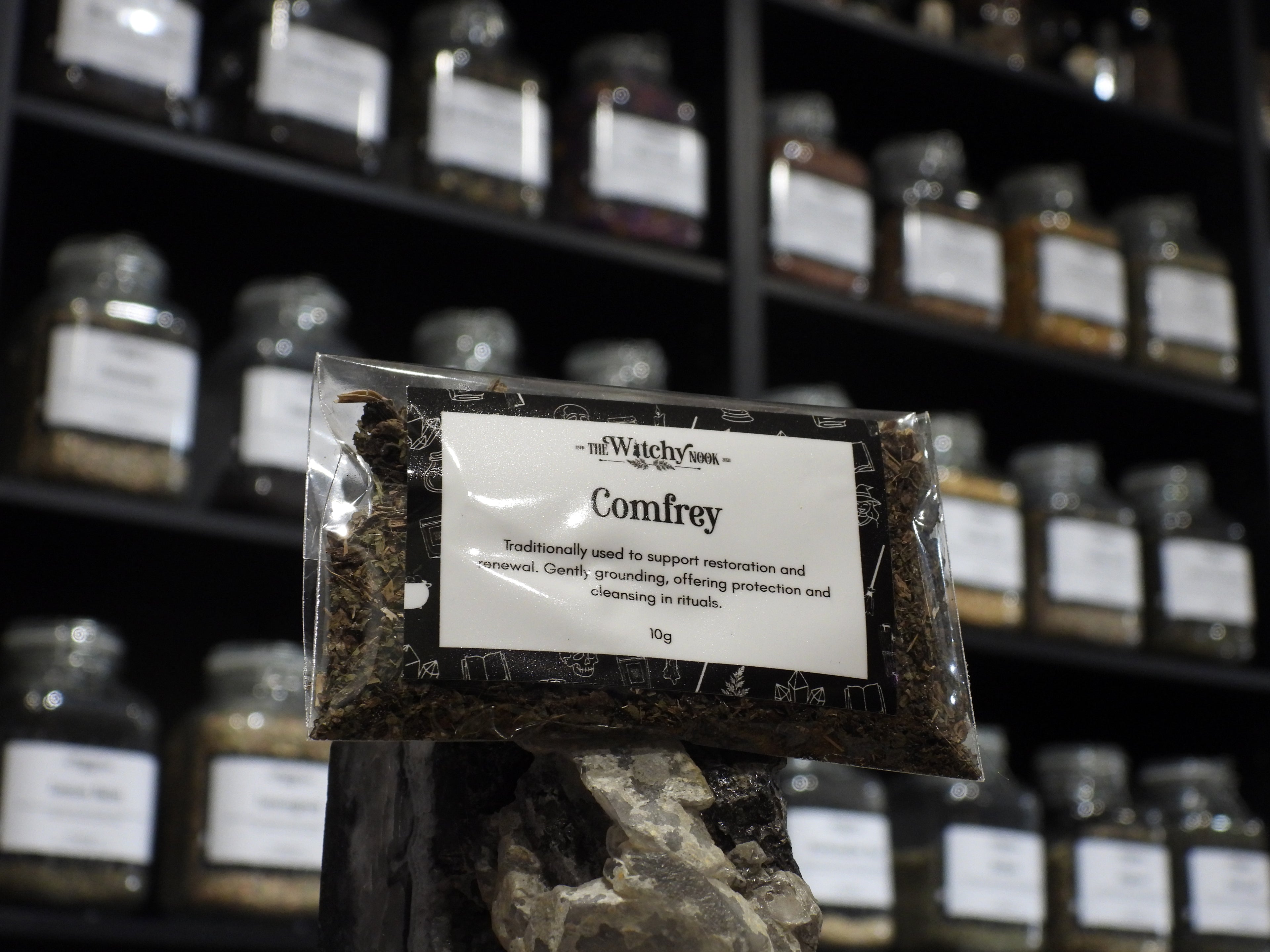 Packaged product labelled ‘Comfrey’ by 'The Witchy Nook' sitting on a crystal with an apothecary of herb jars blurred in the background.