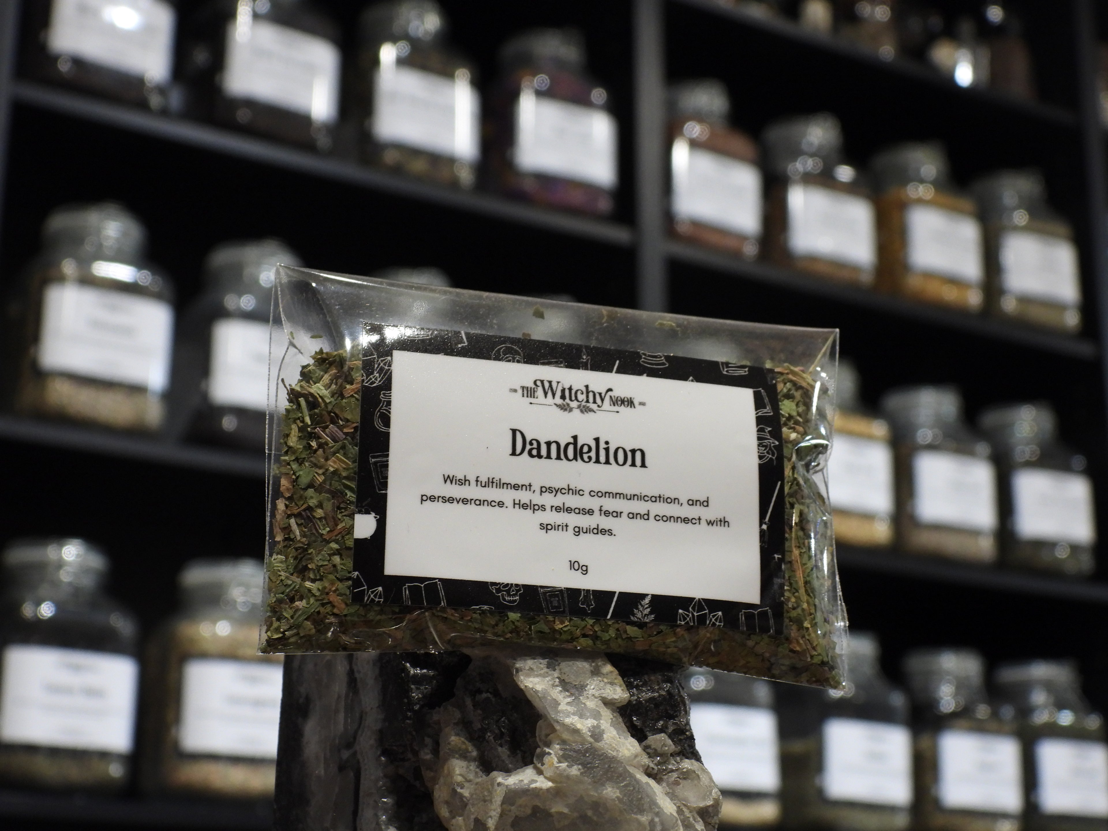 Packaged product labelled ‘Dandelion’ by 'The Witchy Nook' sitting on a crystal with an apothecary of herb jars blurred in the background.