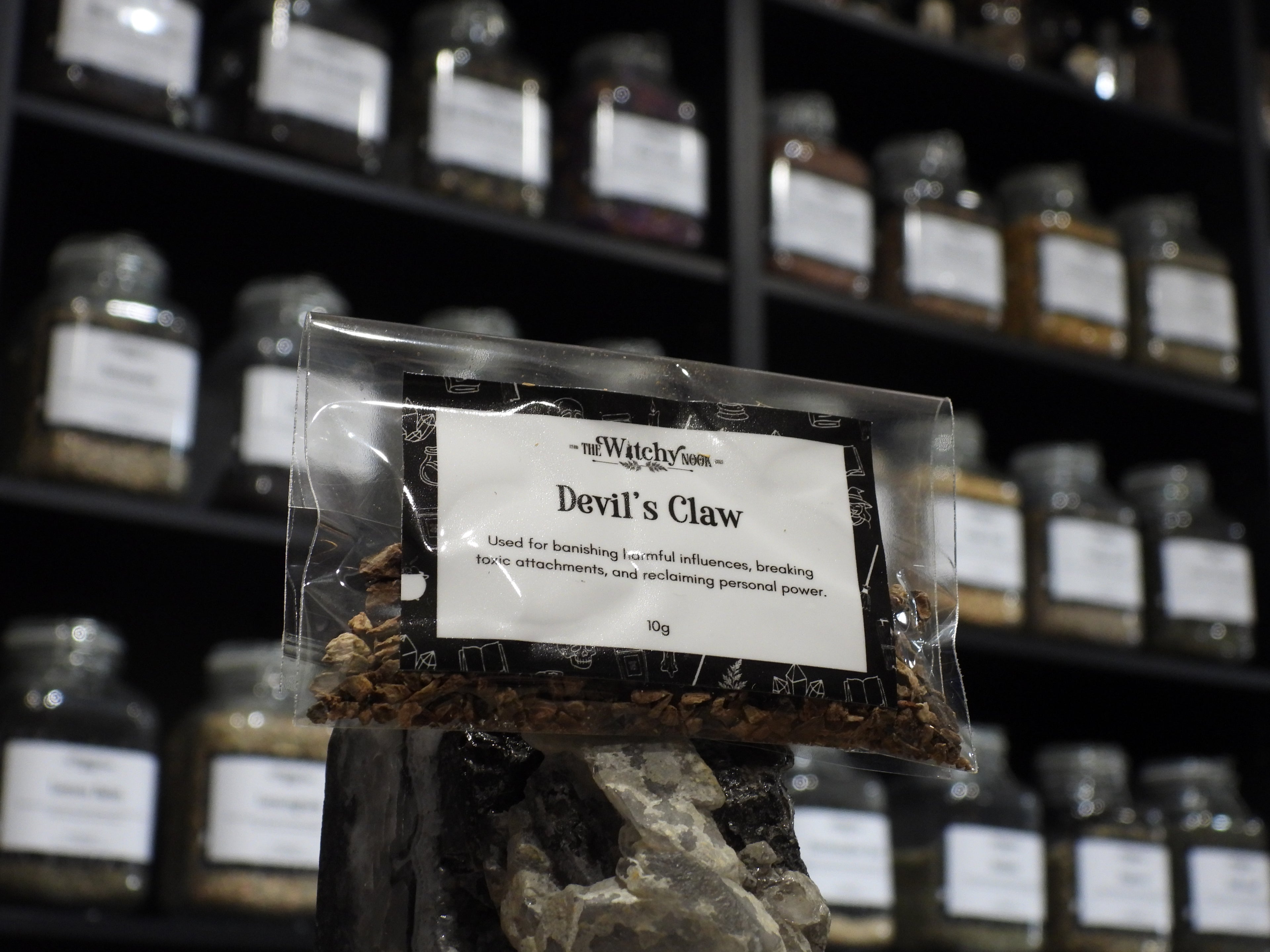 Packaged product labelled ‘Devil’s Claw' by 'Witchy Nook' sitting on a crystal with an apothecary of herb jars blurred in the background.