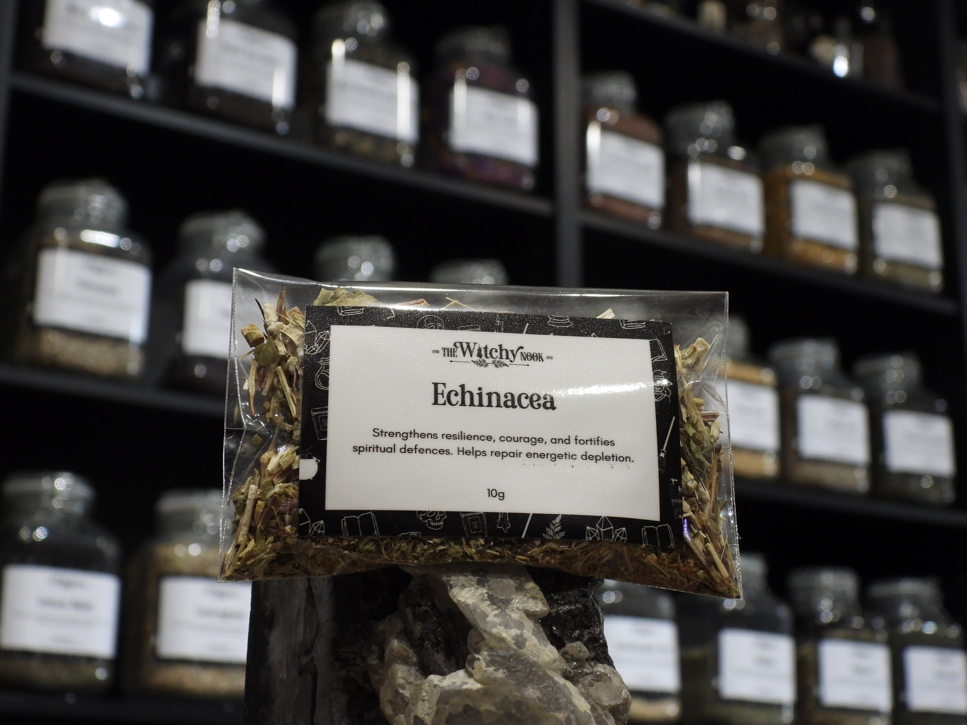 Packaged product labelled ‘Echinacea' by 'Witchy Nook' sitting on a crystal with an apothecary of herb jars blurred in the background.