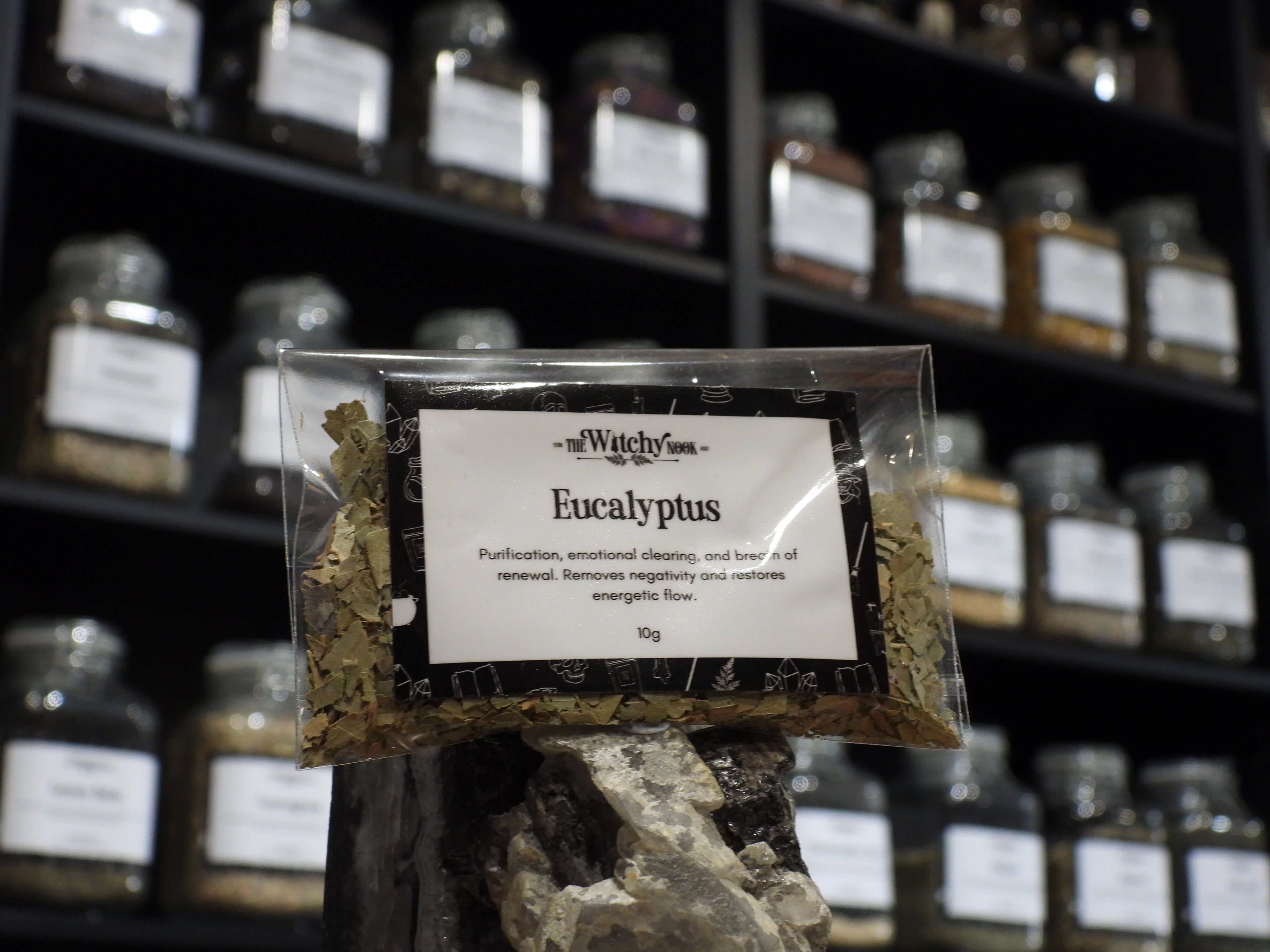 Packaged product labelled ‘Eucalyptus' by 'Witchy Nook' sitting on a crystal with an apothecary of herb jars blurred in the background.