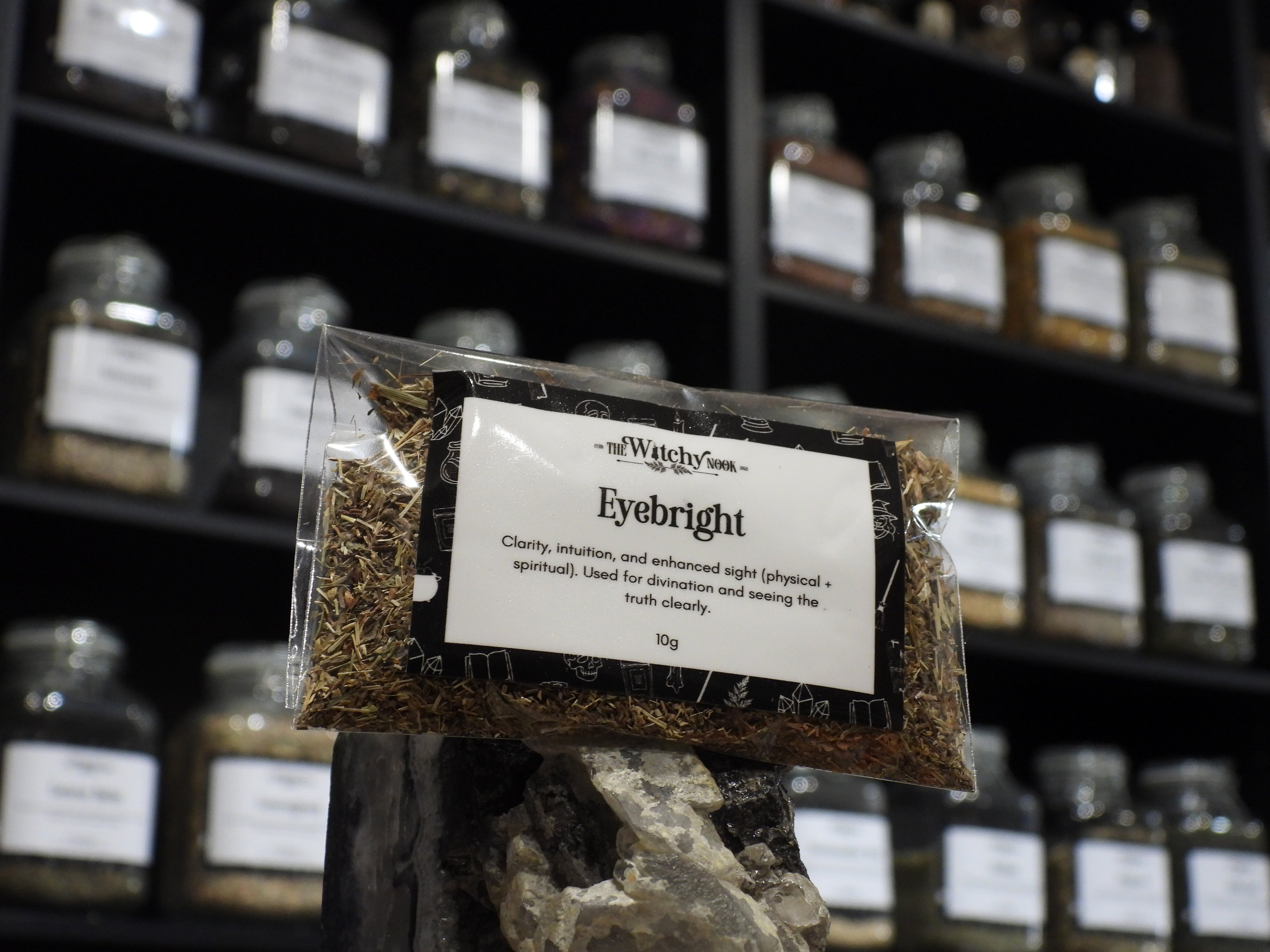 Packaged product labelled ‘Eyebright' by 'Witchy Nook' sitting on a crystal with an apothecary of herb jars blurred in the background.
