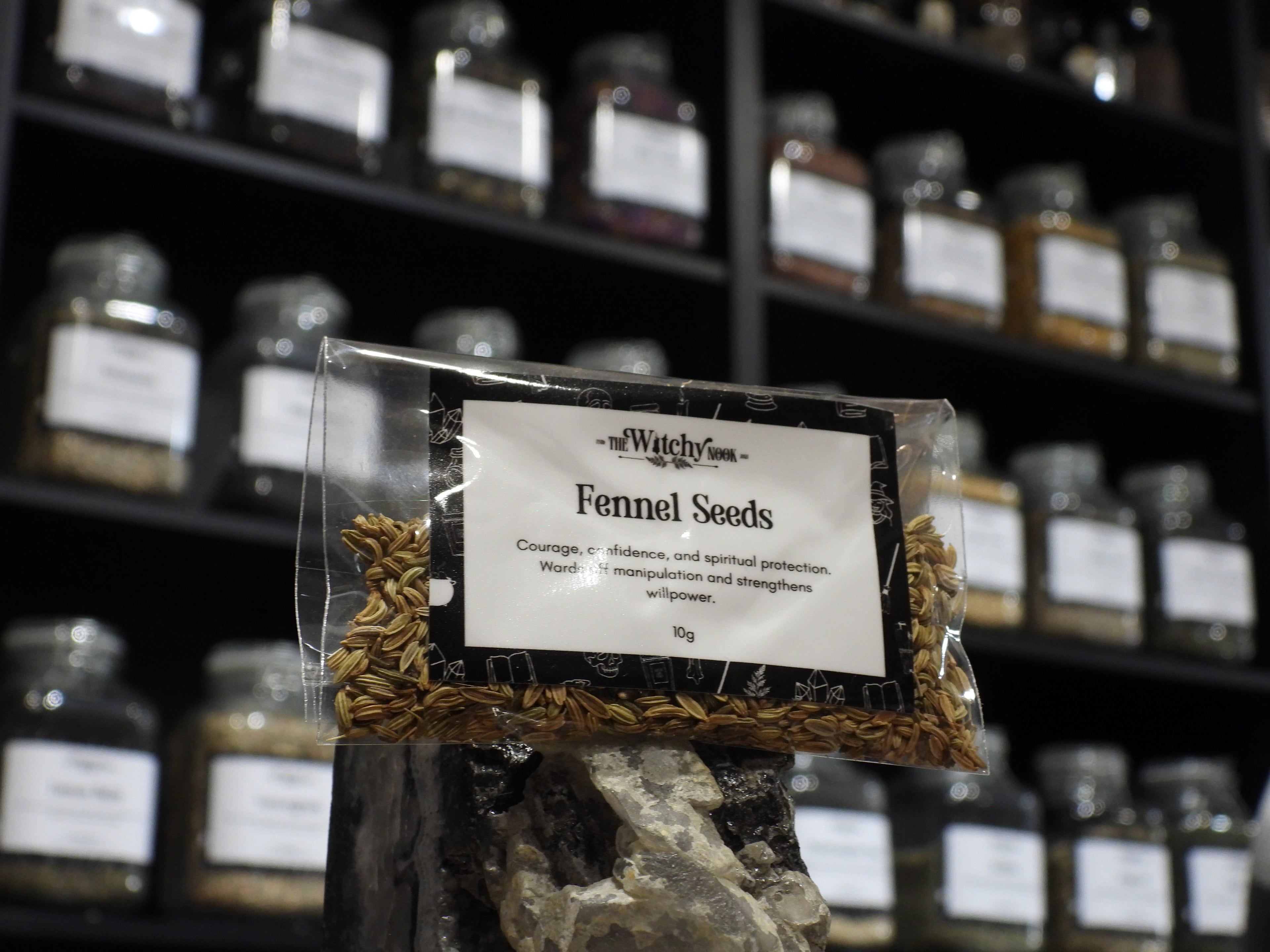 Packaged product labelled ‘Fennel Seeds' by 'Witchy Nook' sitting on a crystal with an apothecary of herb jars blurred in the background.