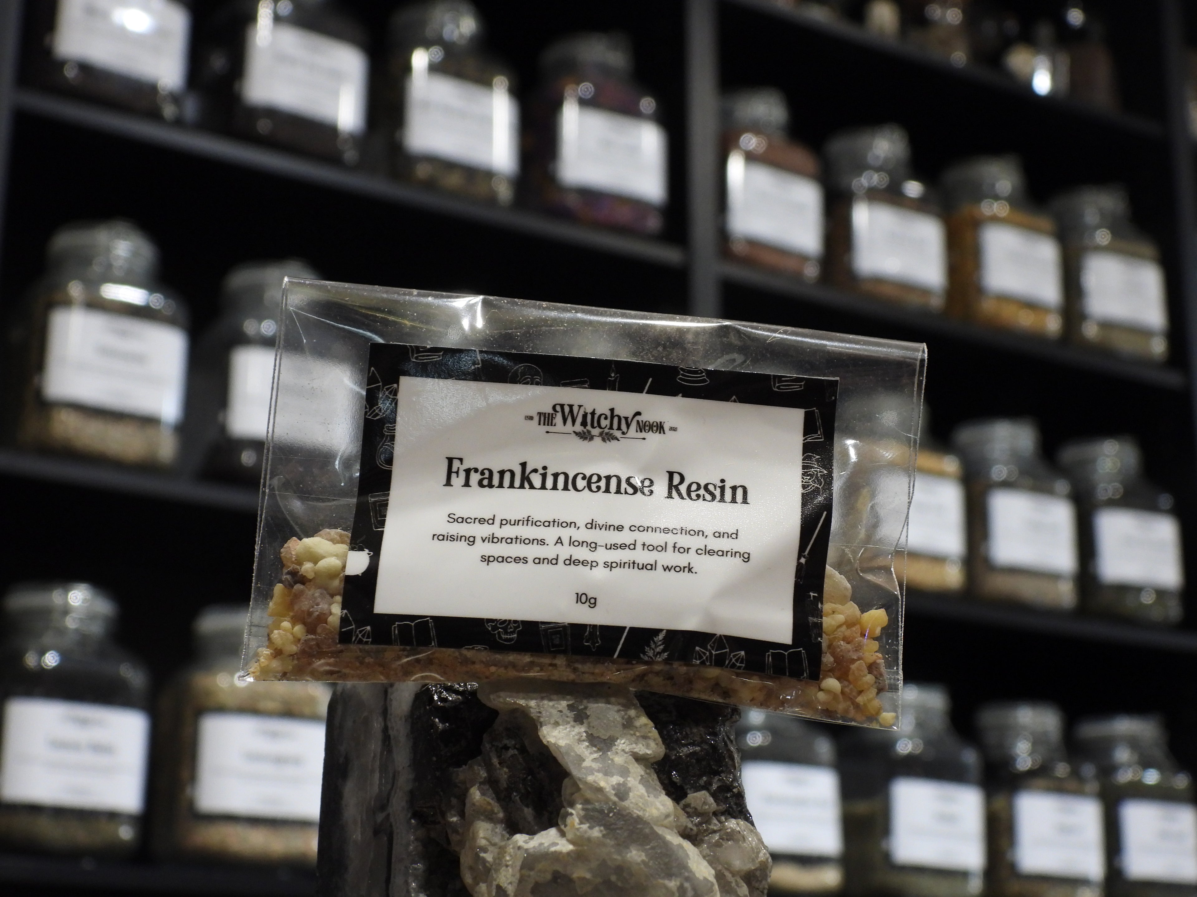 Packaged product labelled ‘Frankincense Resin' by 'Witchy Nook' sitting on a crystal with an apothecary of herb jars blurred in the background.