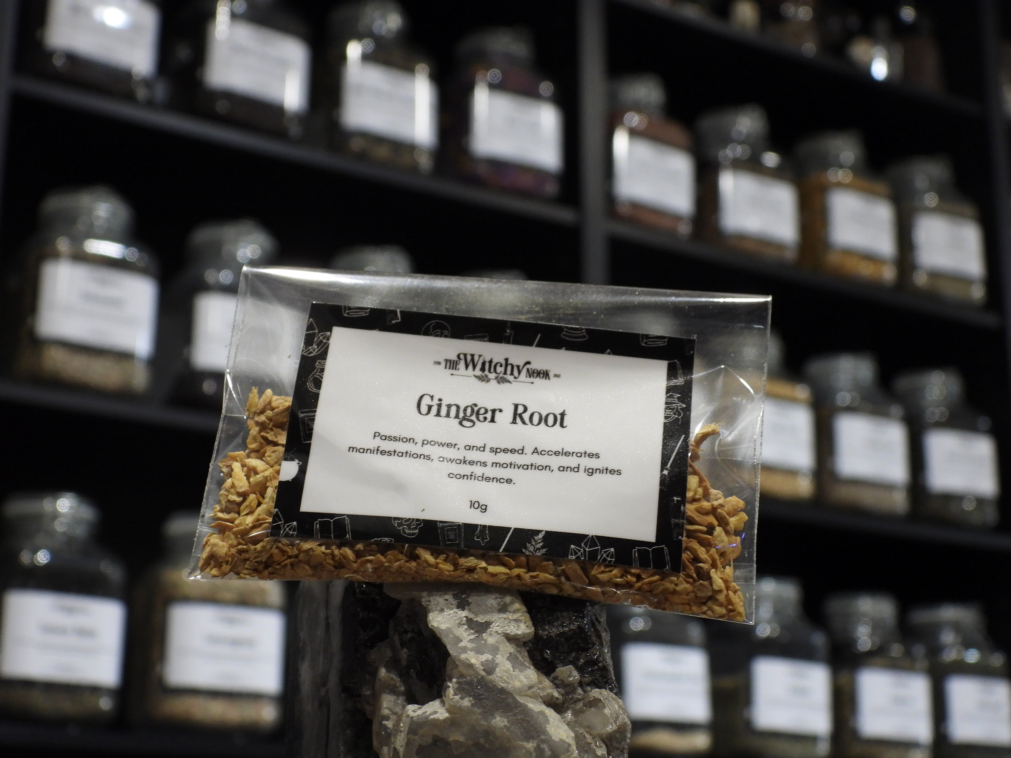 Packaged product labelled ‘Ginger Root' by 'Witchy Nook' sitting on a crystal with an apothecary of herb jars blurred in the background.
