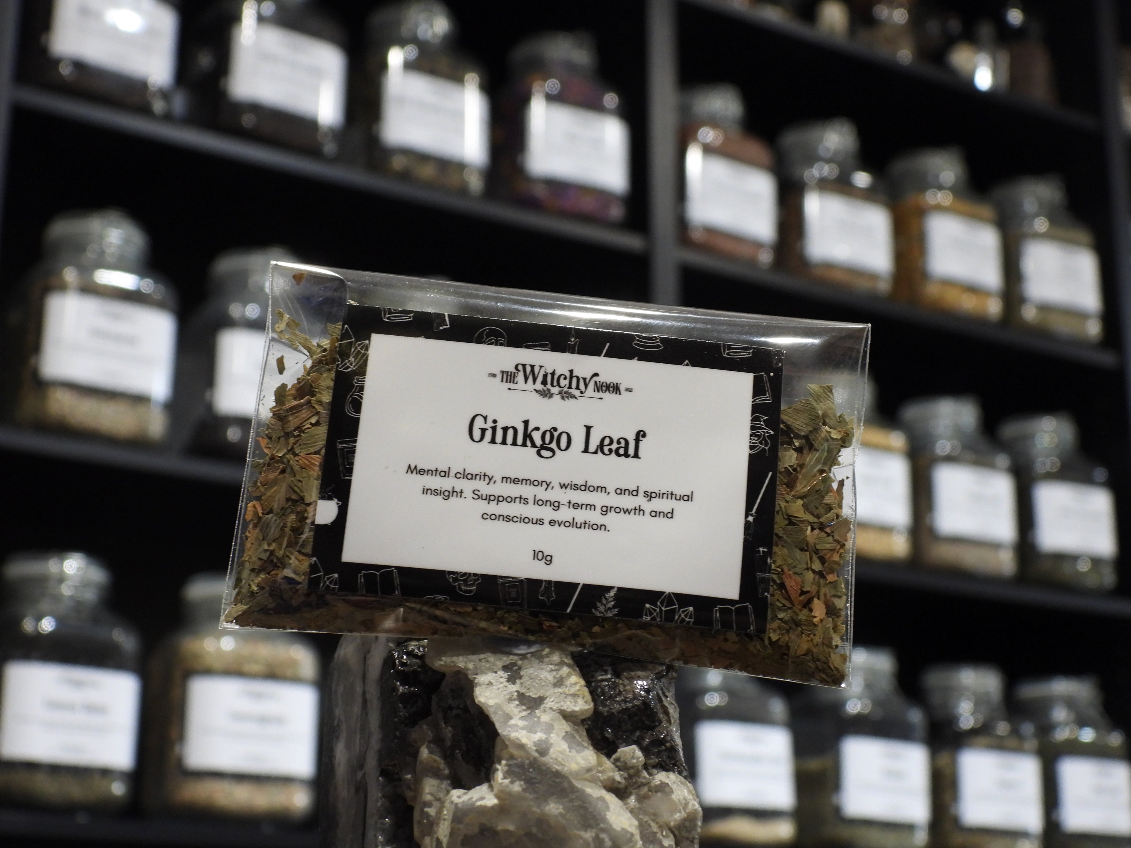 Packaged product labelled ‘Ginkgo Leaf' by 'Witchy Nook' sitting on a crystal with an apothecary of herb jars blurred in the background.