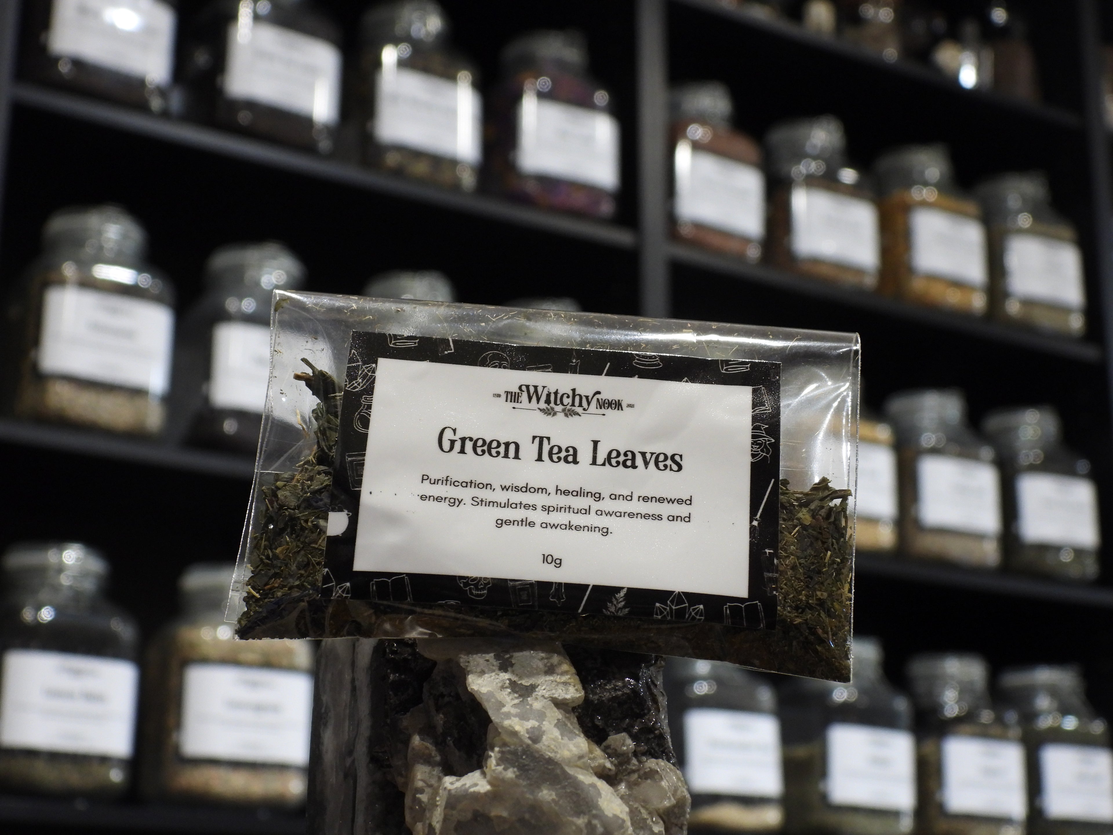 Packaged product labelled ‘Green Tea Leaves' by 'Witchy Nook' sitting on a crystal with an apothecary of herb jars blurred in the background.