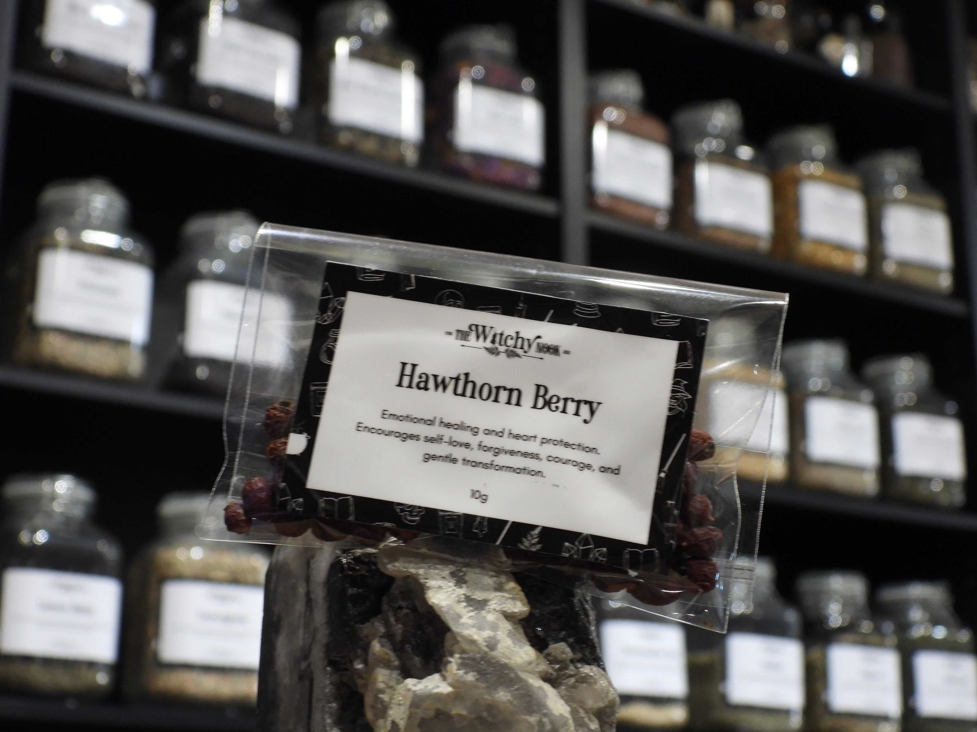 Packaged product labelled ‘Hawthorn Berry’ by 'The Witchy Nook' sitting on a crystal with an apothecary of herb jars blurred in the background.