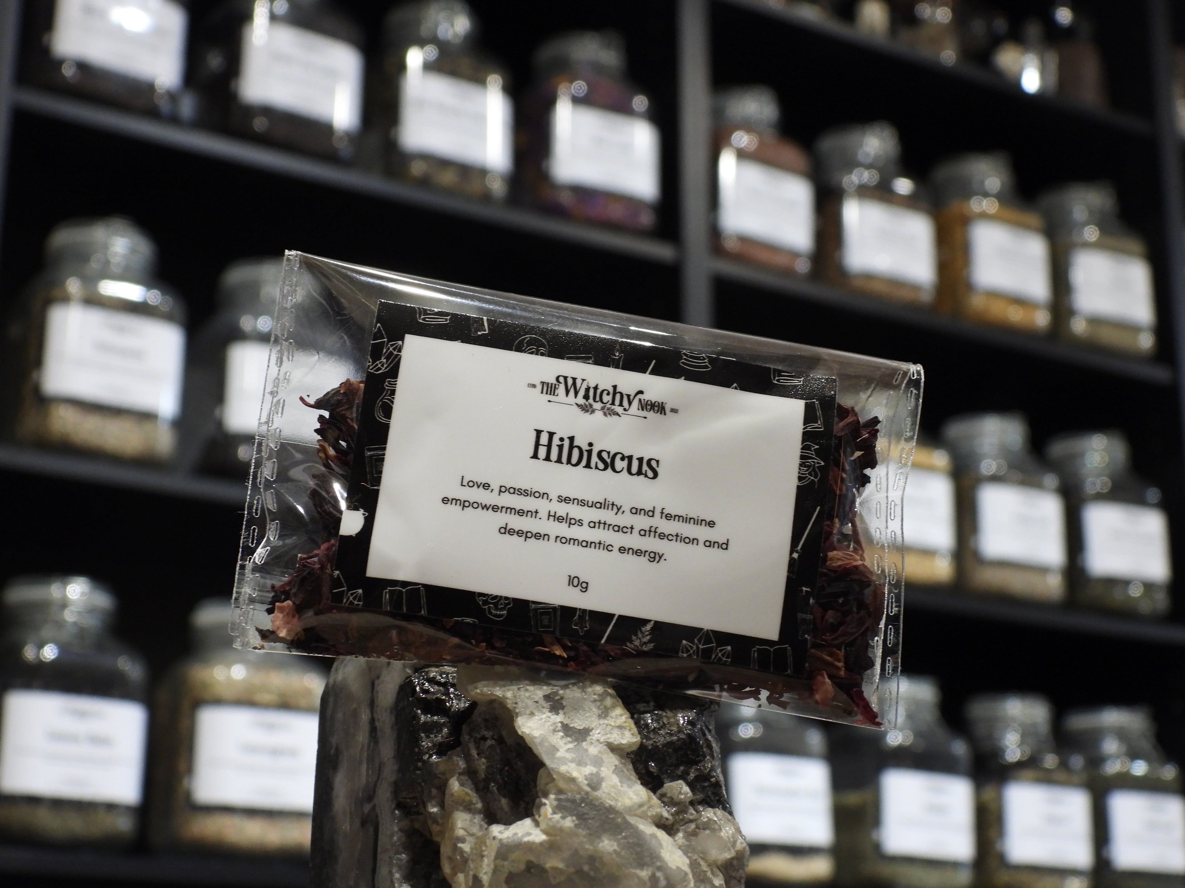 Packaged product labelled ‘Hibiscus’ by 'The Witchy Nook' sitting on a crystal with an apothecary of herb jars blurred in the background.