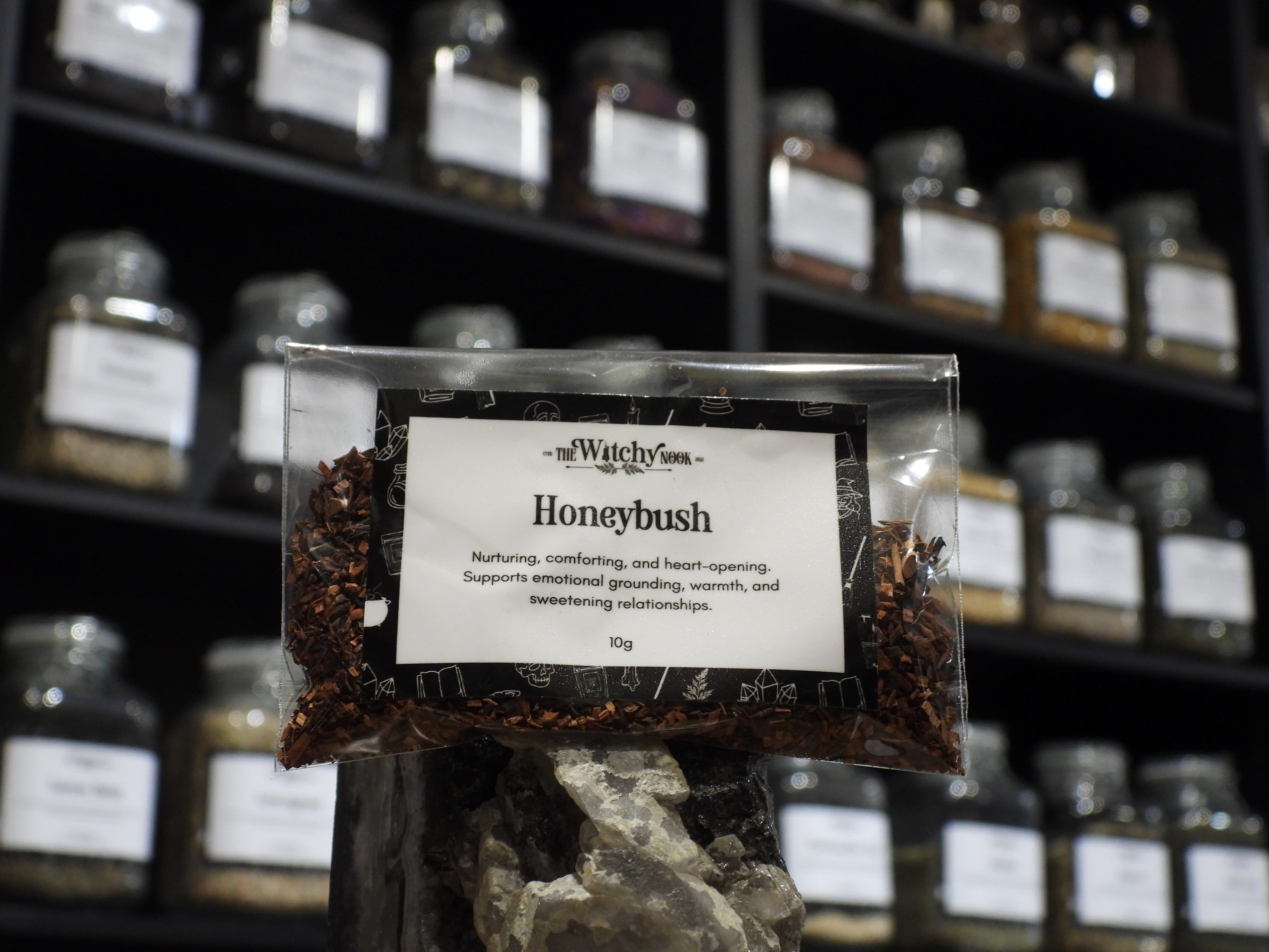 Packaged product labelled ‘Honeybush’ by 'The Witchy Nook' sitting on a crystal with an apothecary of herb jars blurred in the background.