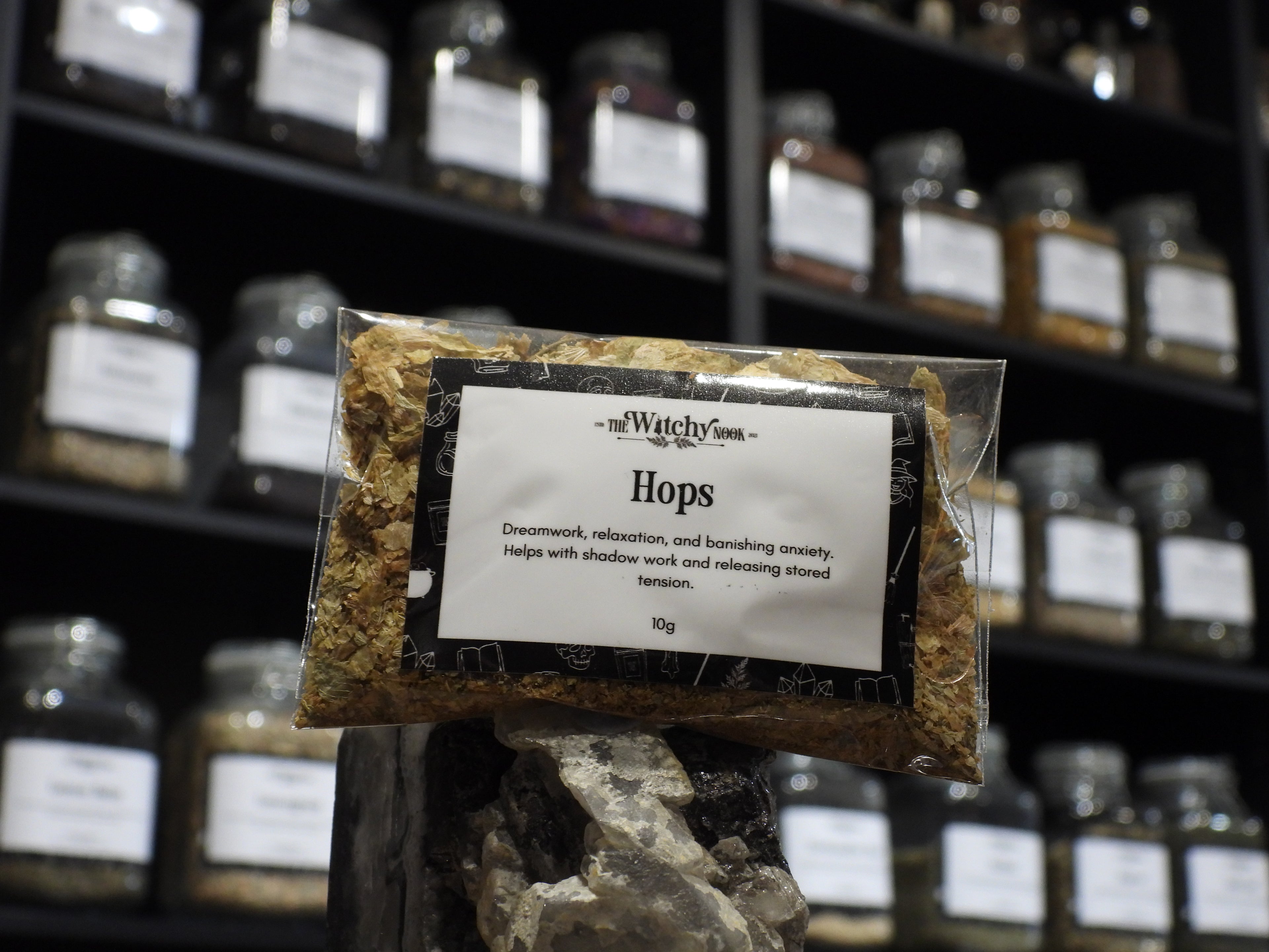 Packaged product labelled ‘Hops' by 'Witchy Nook' sitting on a crystal with an apothecary of herb jars blurred in the background.