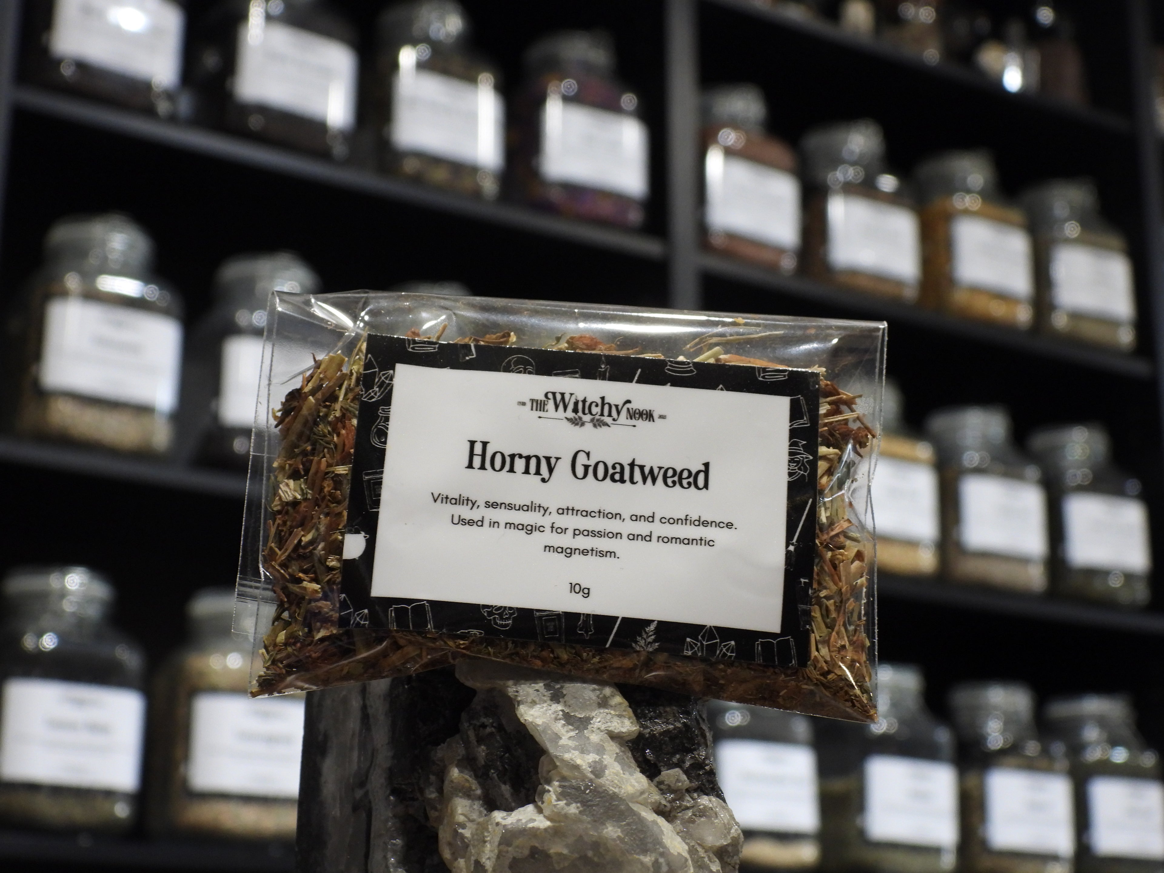 Packaged product labelled ‘Horny Goatweed' by 'Witchy Nook' sitting on a crystal with an apothecary of herb jars blurred in the background.