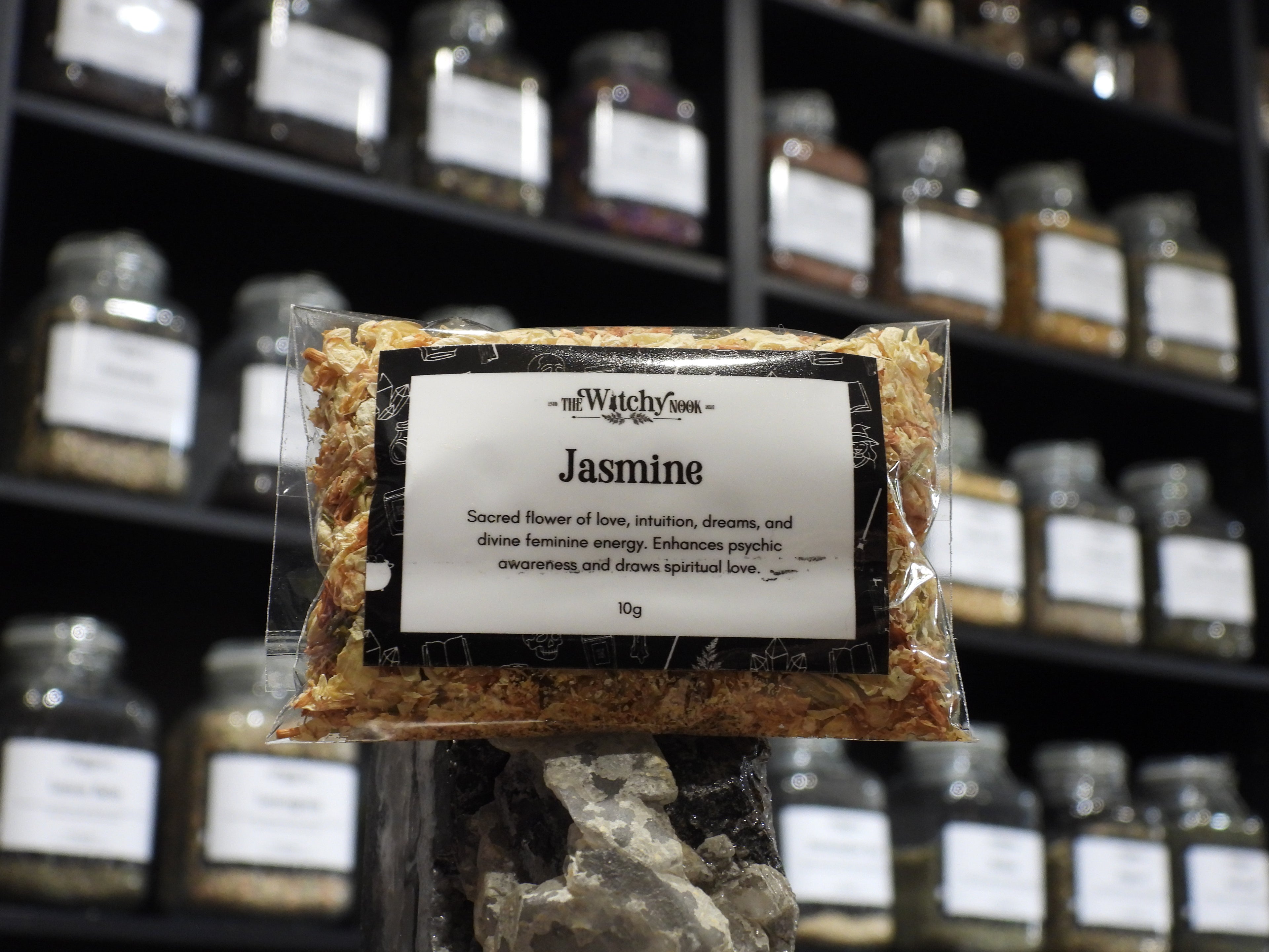 Packaged product labelled ‘Jasmine' by 'Witchy Nook' sitting on a crystal with an apothecary of herb jars blurred in the background.